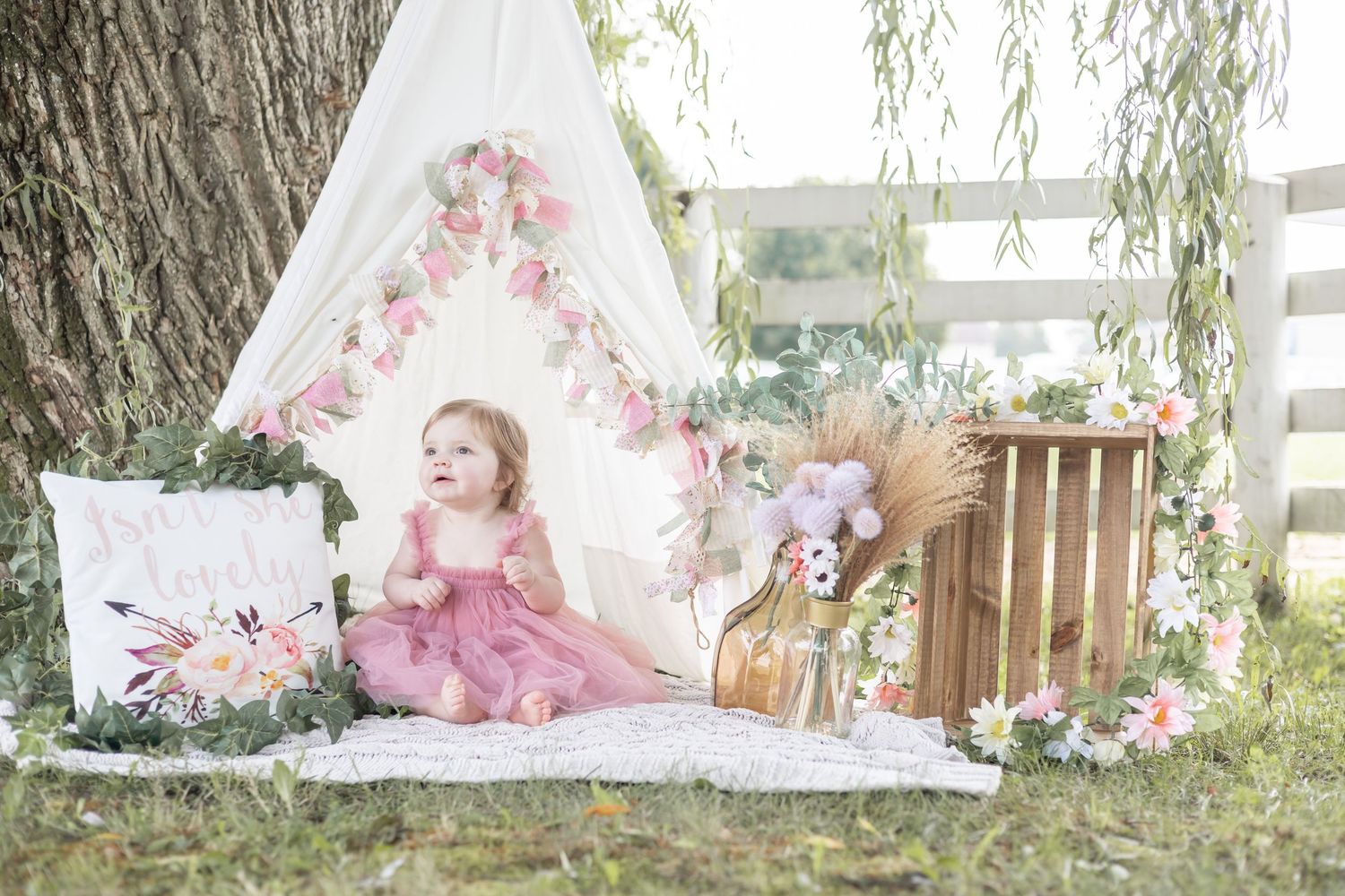 A whimsical outdoor photo session setup with white teepee tents, floral decorations and rustic wooden elements in a garden.