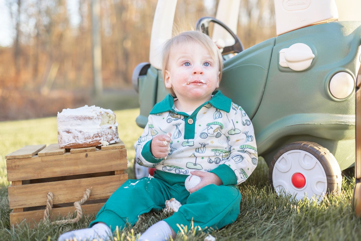 Small child in spring green overalls sits on grass next to vintage pedal car and wooden crate in outdoor setting.