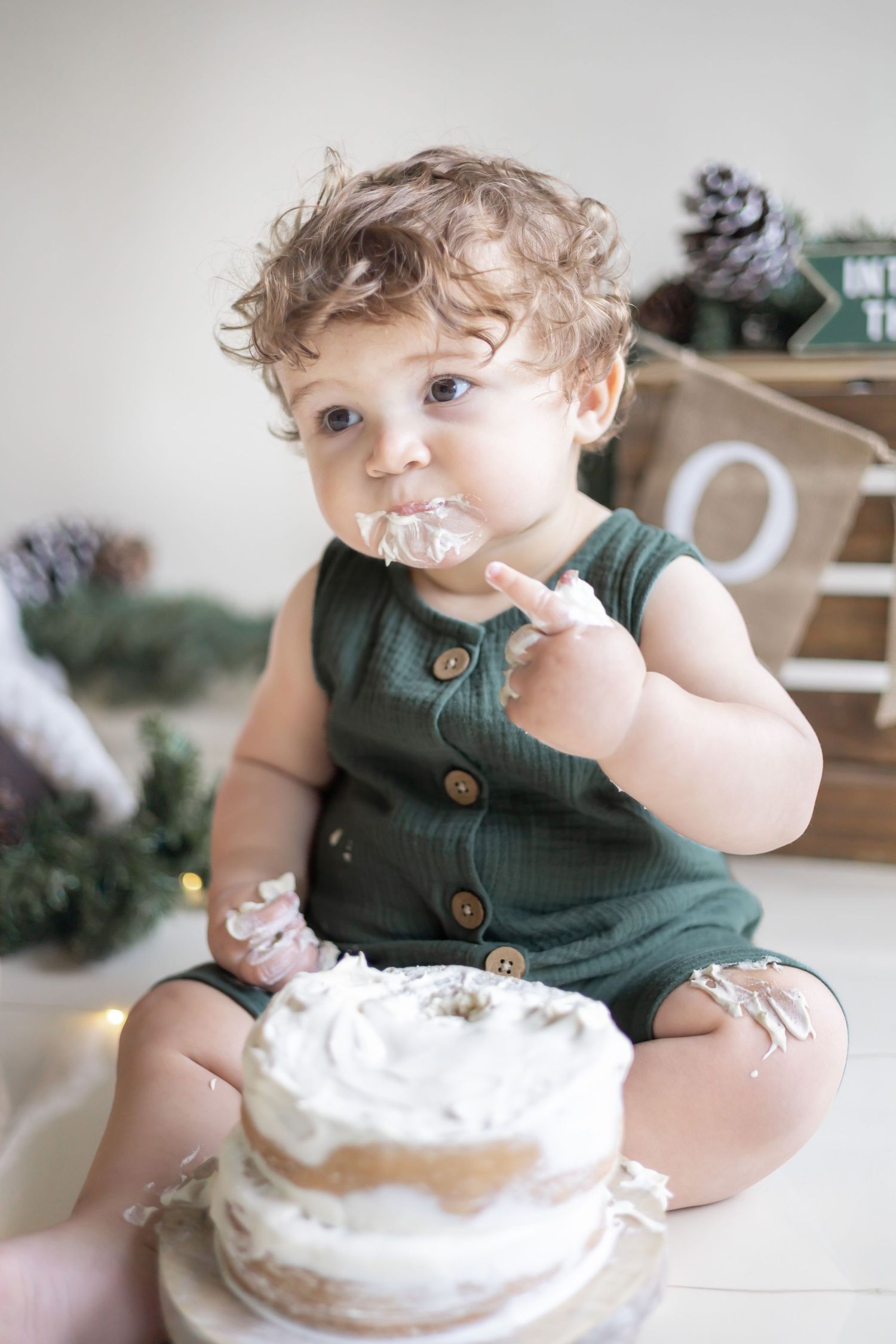 Small child in green overalls sits on floor messily eating white frosted cake during cake smash photo session.