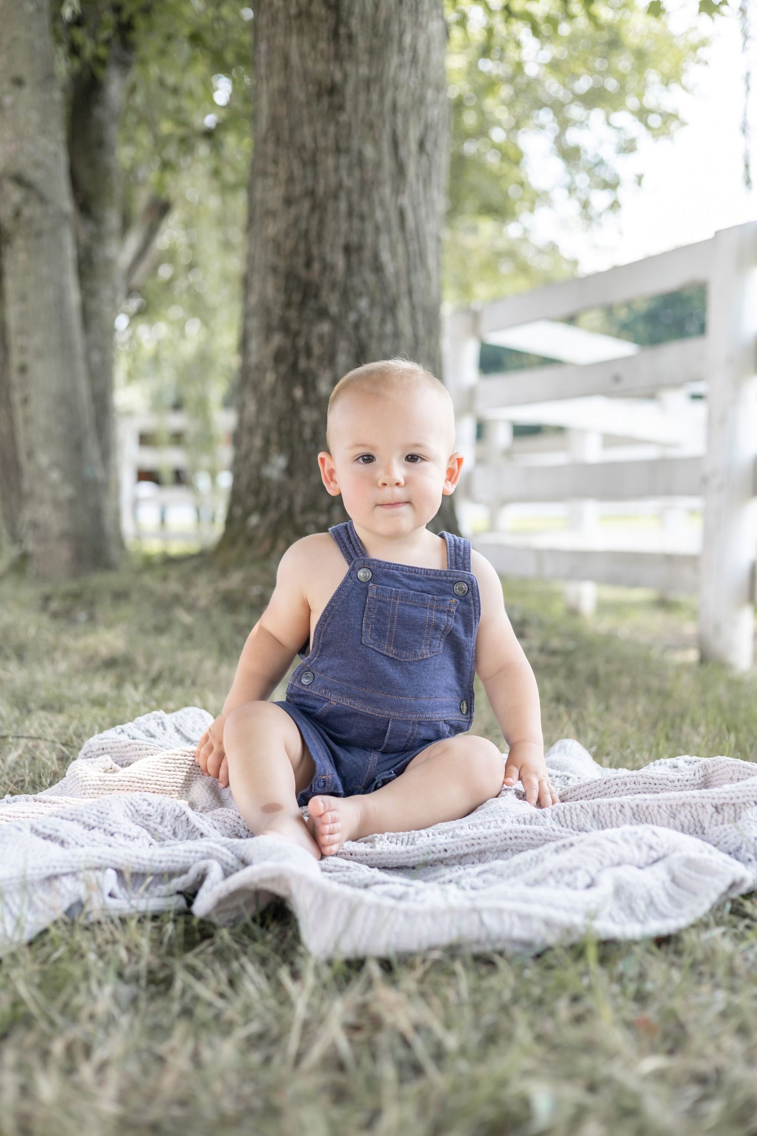 Baby in blue overalls sits on white blanket under shady trees in outdoor photo shoot.