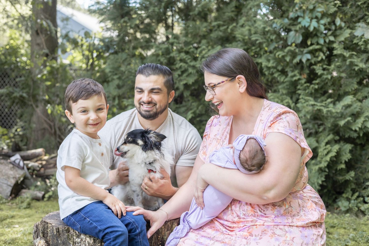 A family sitting together and laughing outdoors in a wooded area during a summer photo session.
