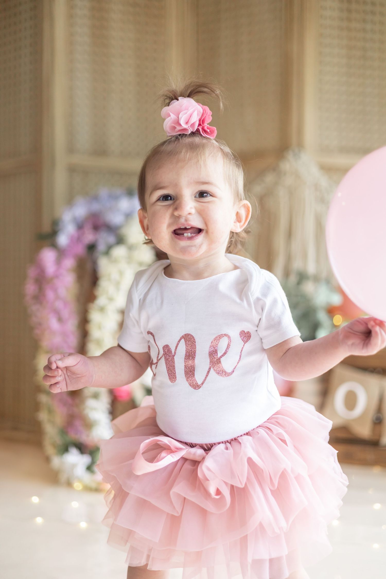 Happy toddler wearing a white onesie and pink tutu dress poses with floral decor in the background.
