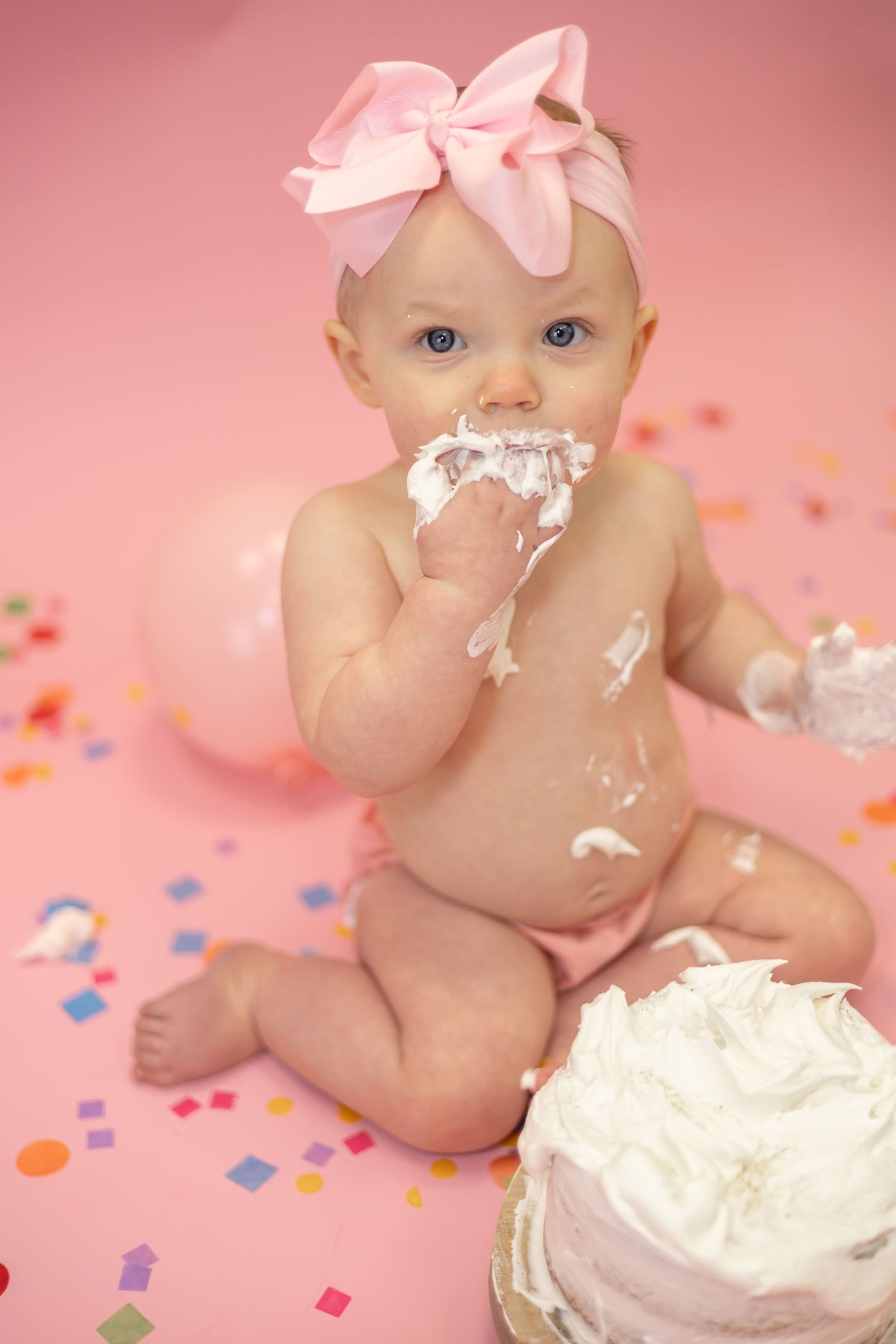 Baby in pink bow enjoying cake smash photo session with white frosting and colorful confetti on pink background.