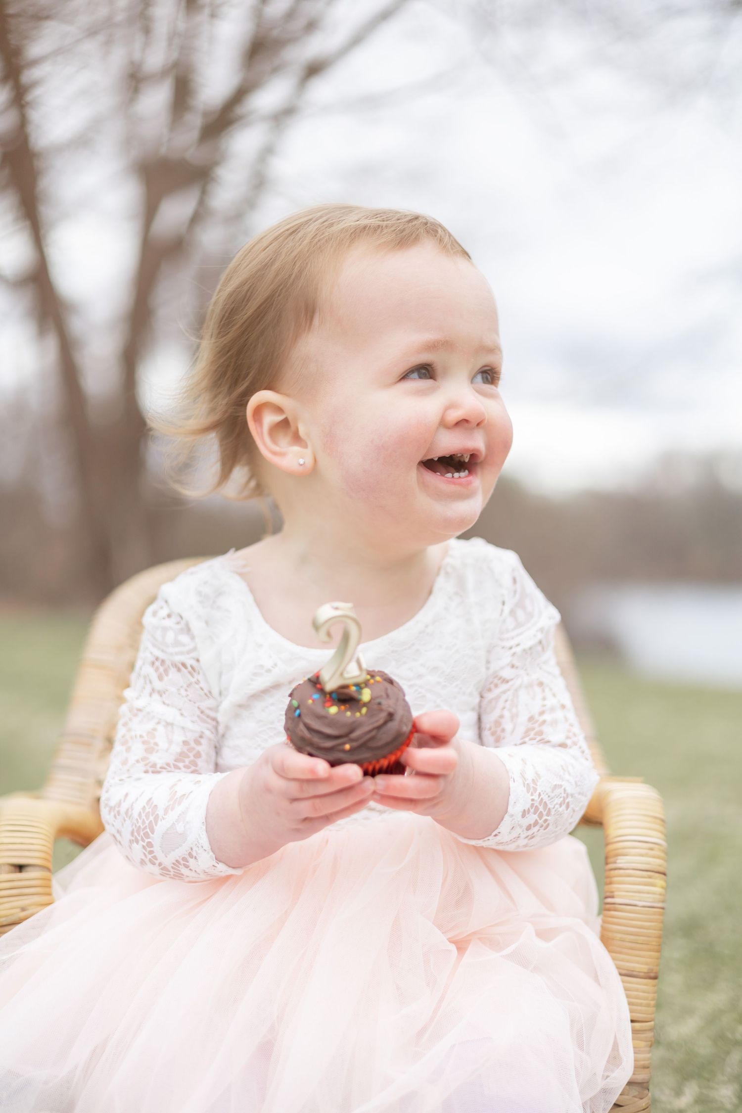 A small child in a pink lace dress sits outdoors enjoying chocolate donuts while smiling and laughing.