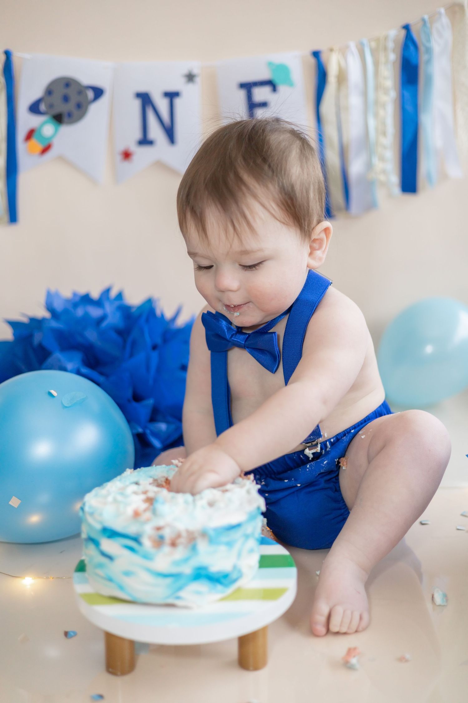 Baby in blue bow tie outfit reaching for birthday cake with blue decorations and balloons at celebration photoshoot.