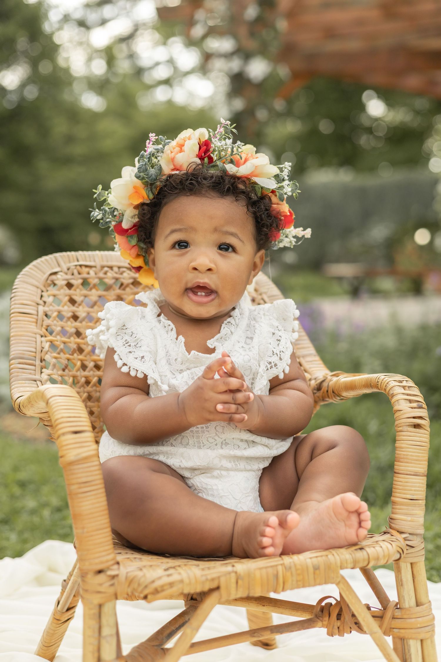 Baby wearing white lace dress and floral crown sits in rattan chair during outdoor photo session.