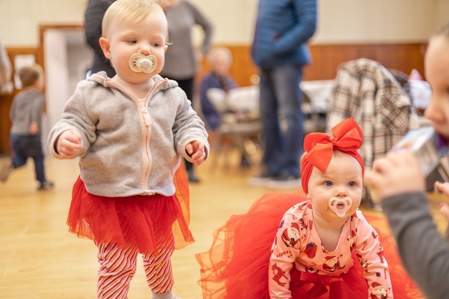 Two small children in red outfits dance and play together on a wooden gymnasium floor during an indoor event.