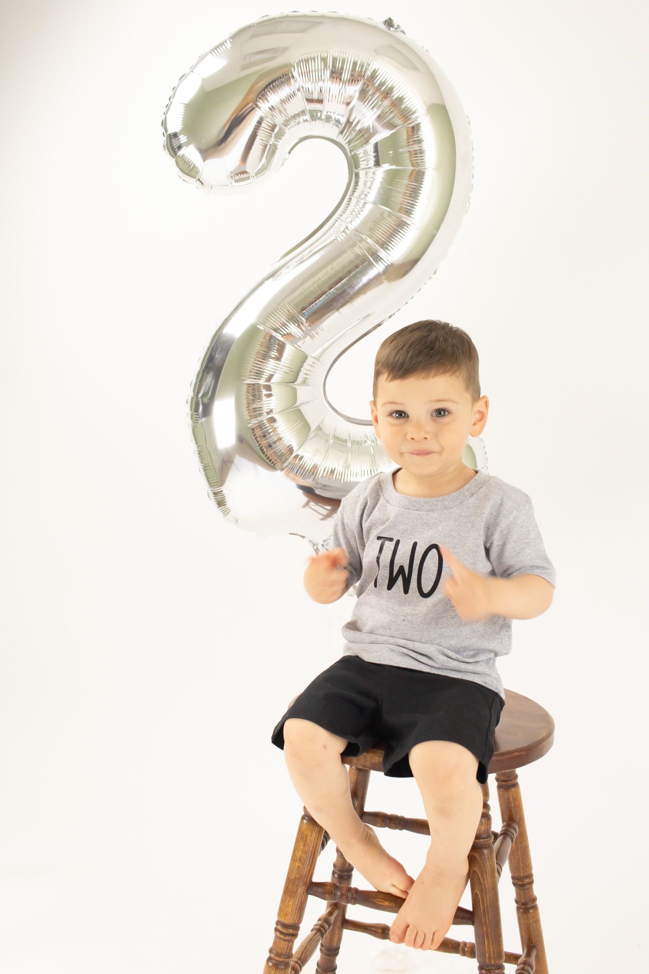 Toddler in gray t-shirt sits on wooden stool with silver number 2 balloon for birthday photo shoot.