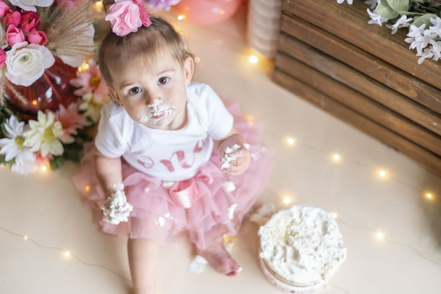 Young children in white and pink outfits surrounded by pink and white floral decorations and twinkling lights.
