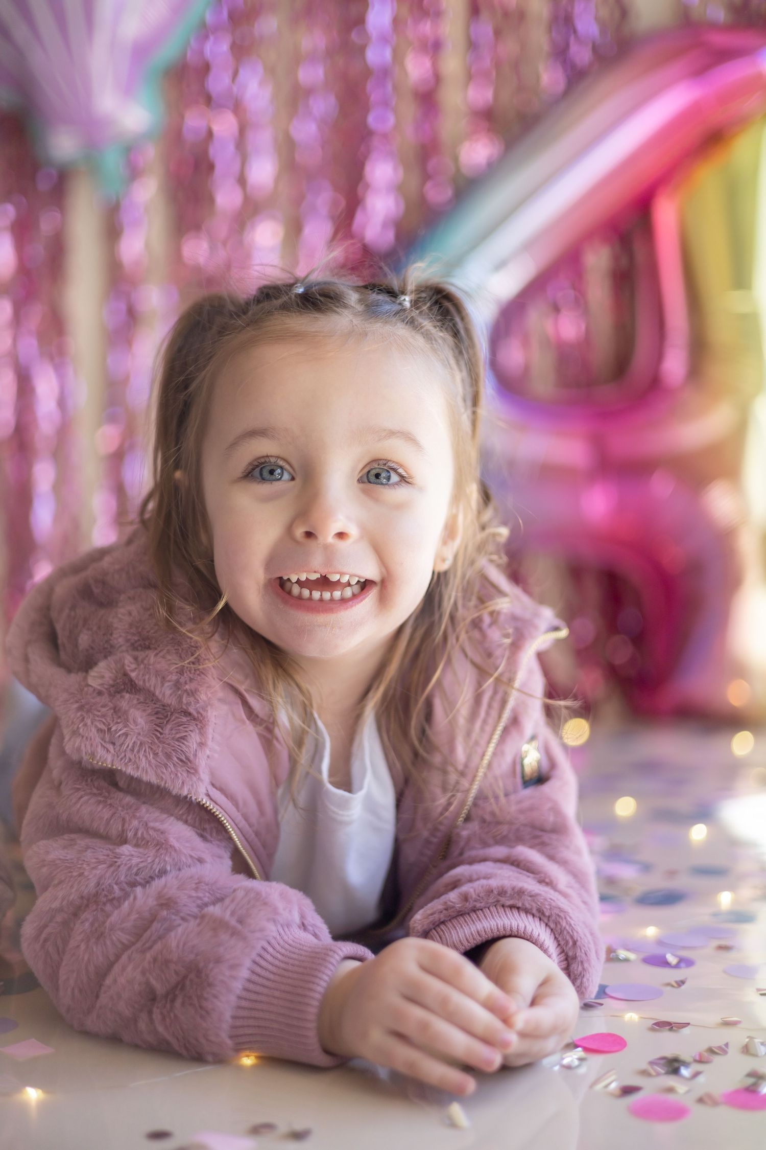 Whimsical birthday scene with pink fuzzy jacket against sparkly backdrop with colorful balloons and confetti.