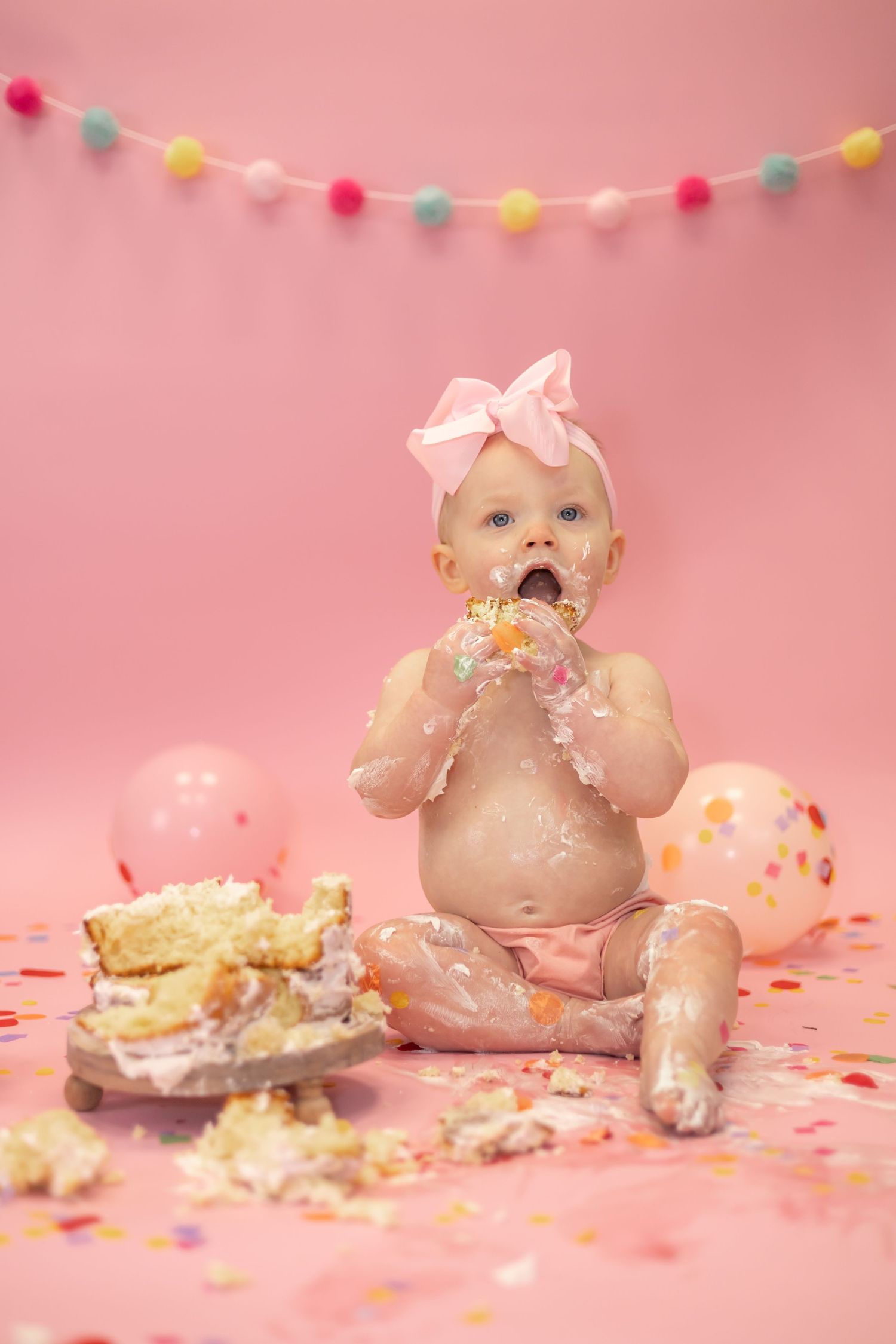 Small child enjoys a birthday cake smash photoshoot with pink decorations and confetti on pink background.