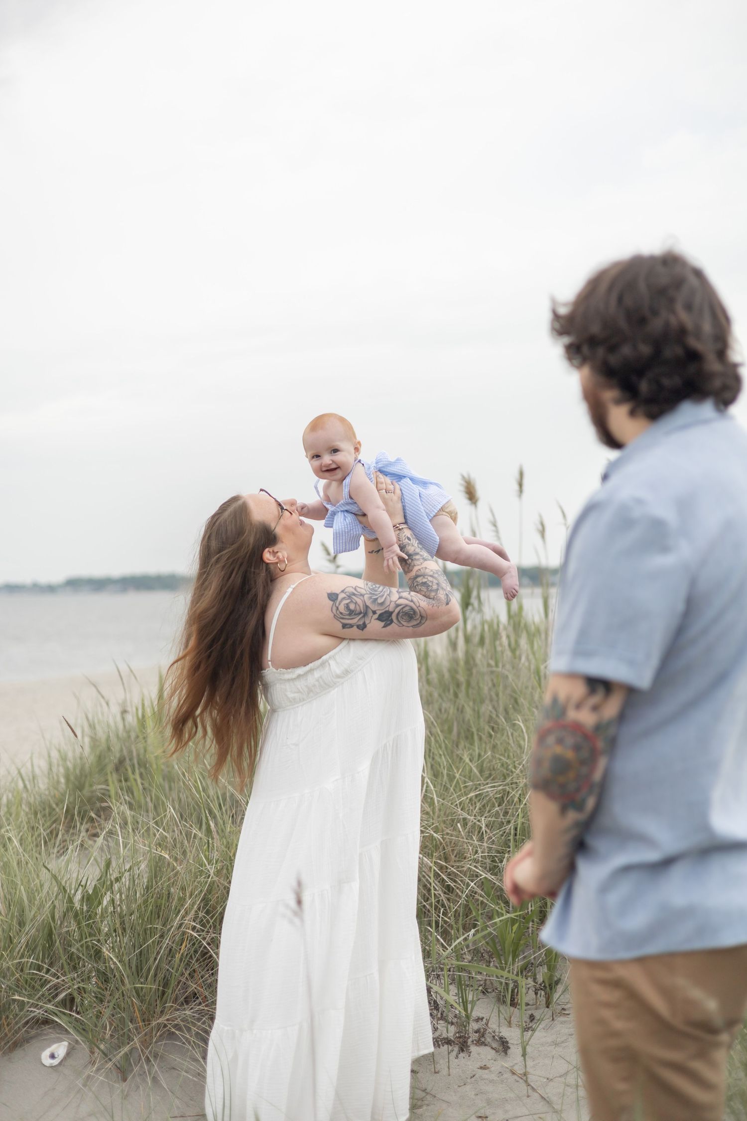 A family shares a tender moment on a beach with tall grass, waves and overcast sky in the background.