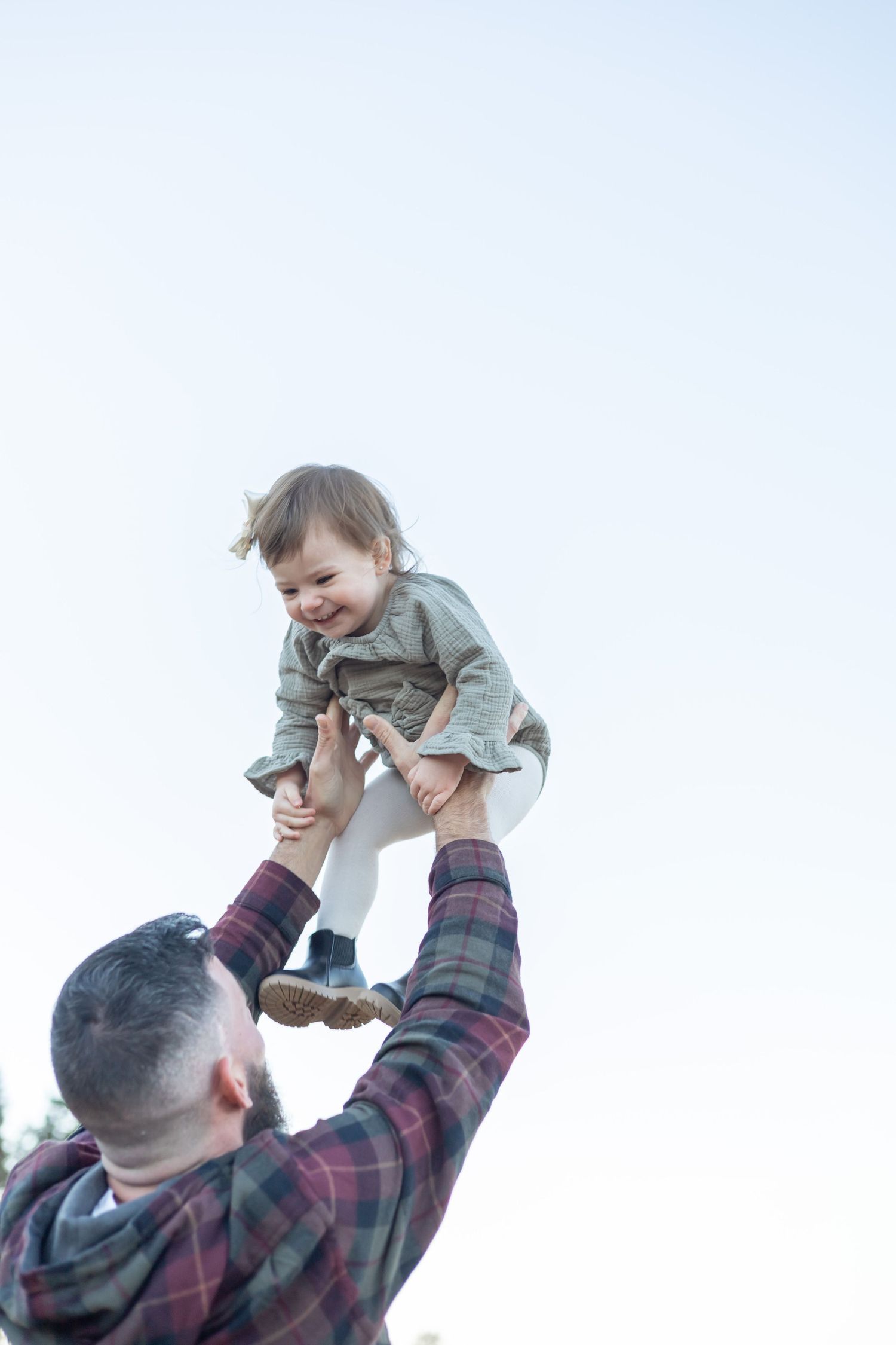 Adult in plaid shirt lifts and plays with a laughing child wearing a gray jacket.