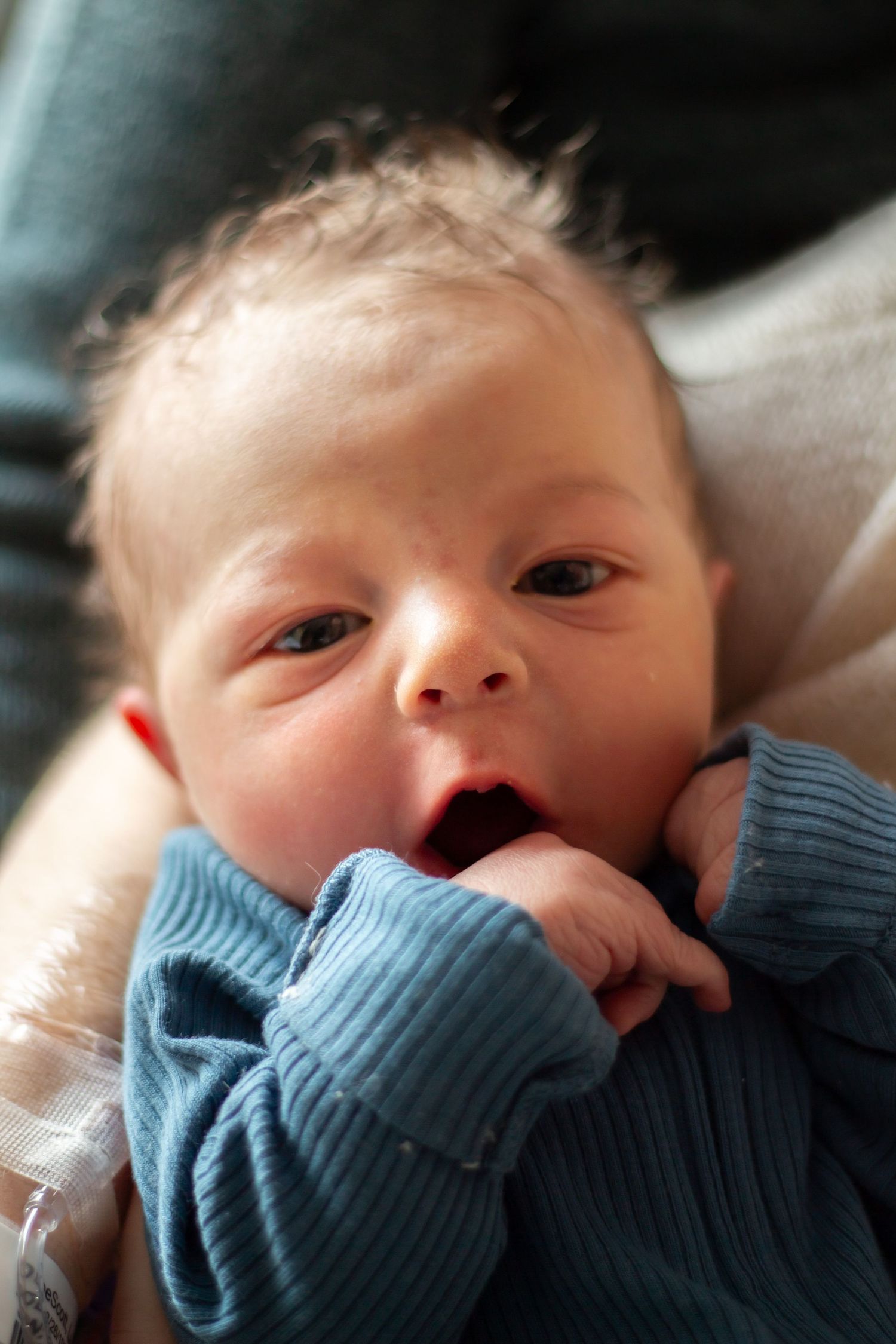 Baby in light blue ribbed sweater lying down with hands near mouth showing curious expression.