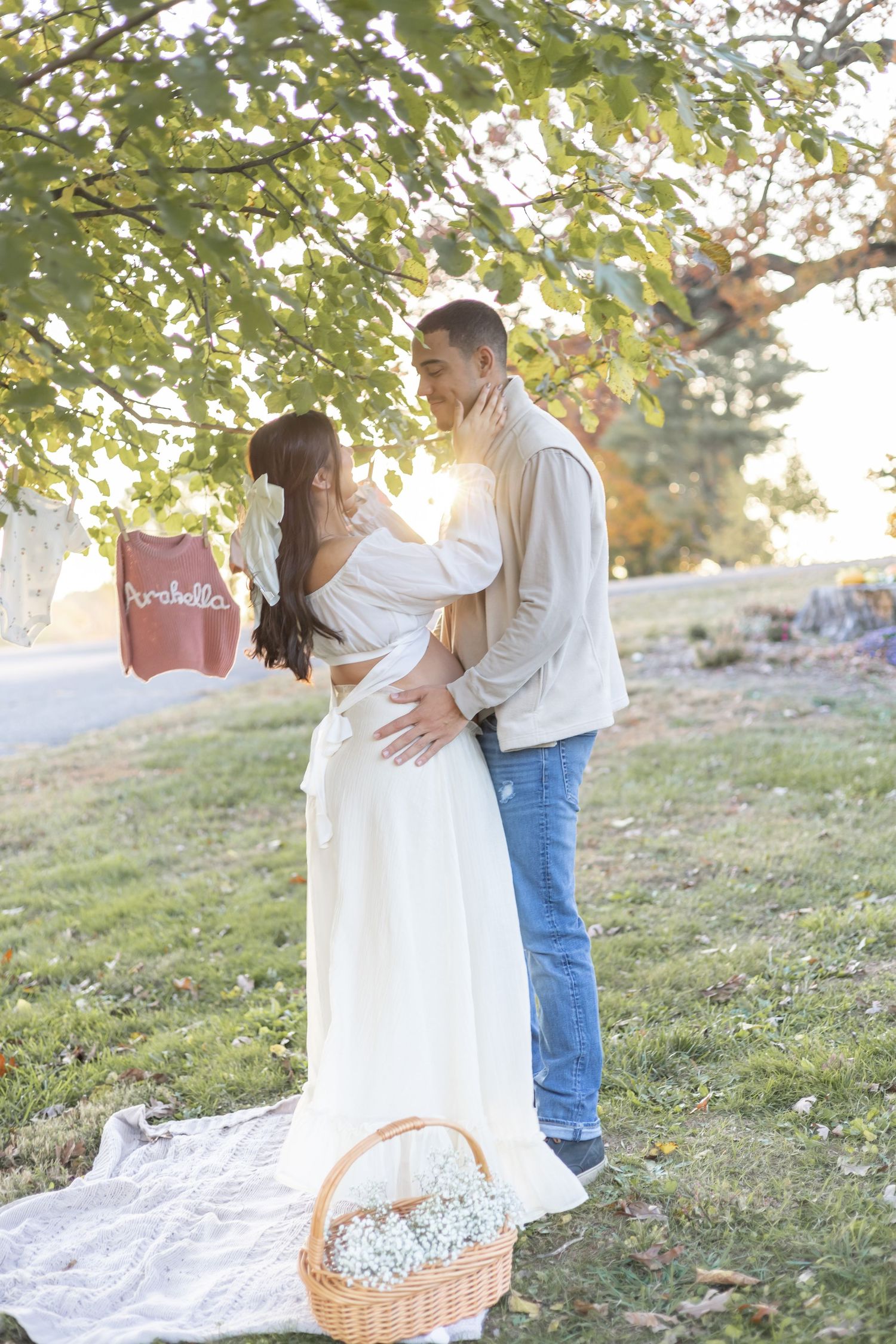 A romantic maternity photoshoot outdoors with a white picnic blanket and wicker basket under autumn foliage at sunset.