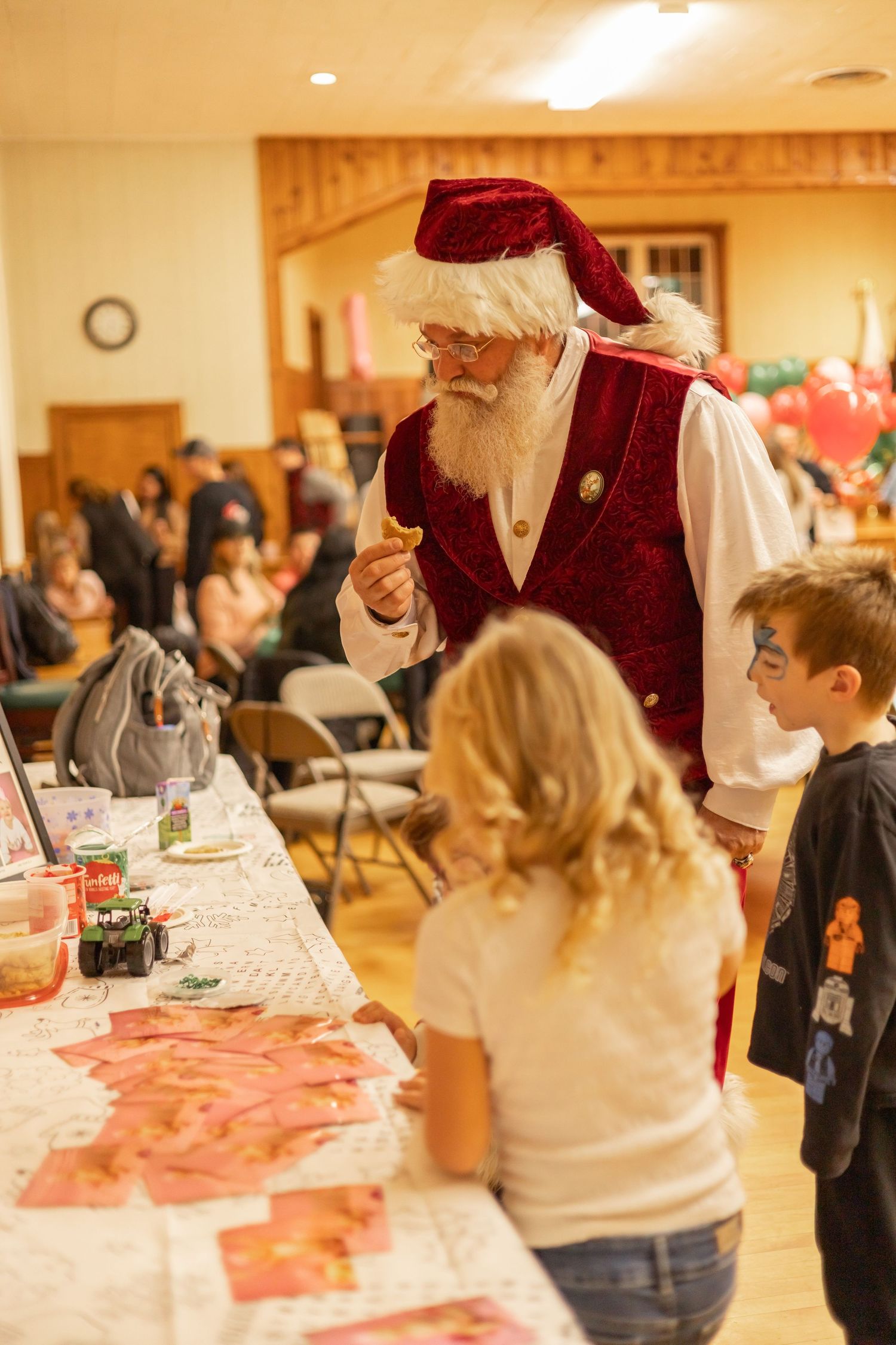 People gather at a warm-lit indoor holiday party with festive decorations and long tables arranged for communal dining.