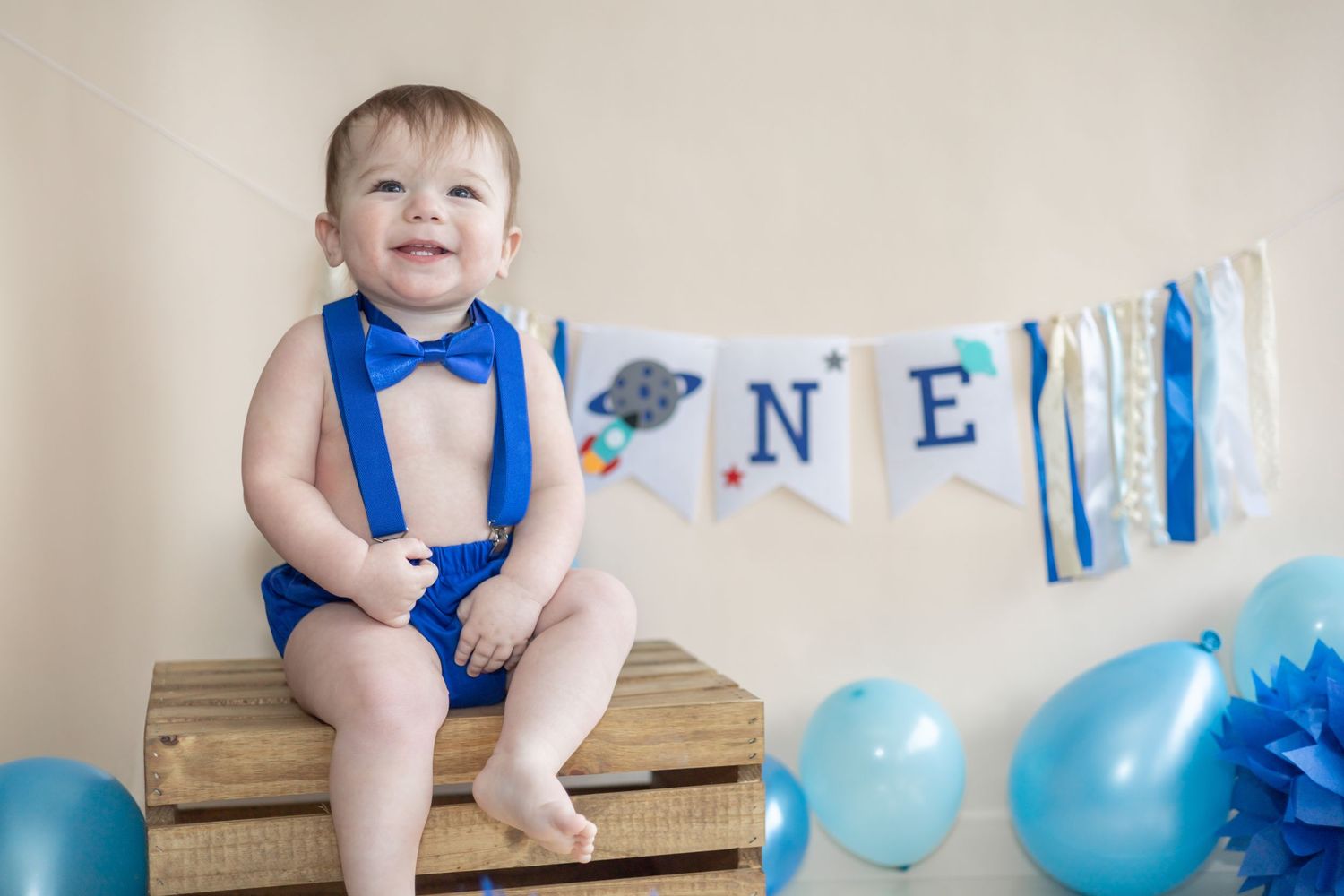 A baby in blue sits on a wooden bench with blue balloons decorating the rustic indoor party scene.