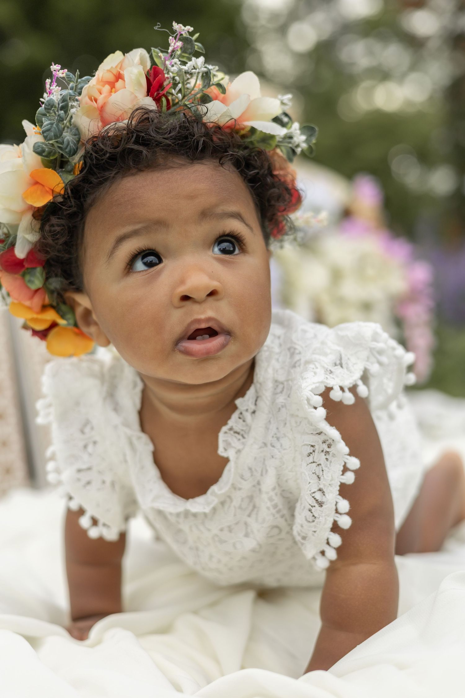 Baby wearing white lace dress and floral crown gazes upward while lying on blanket in garden setting.