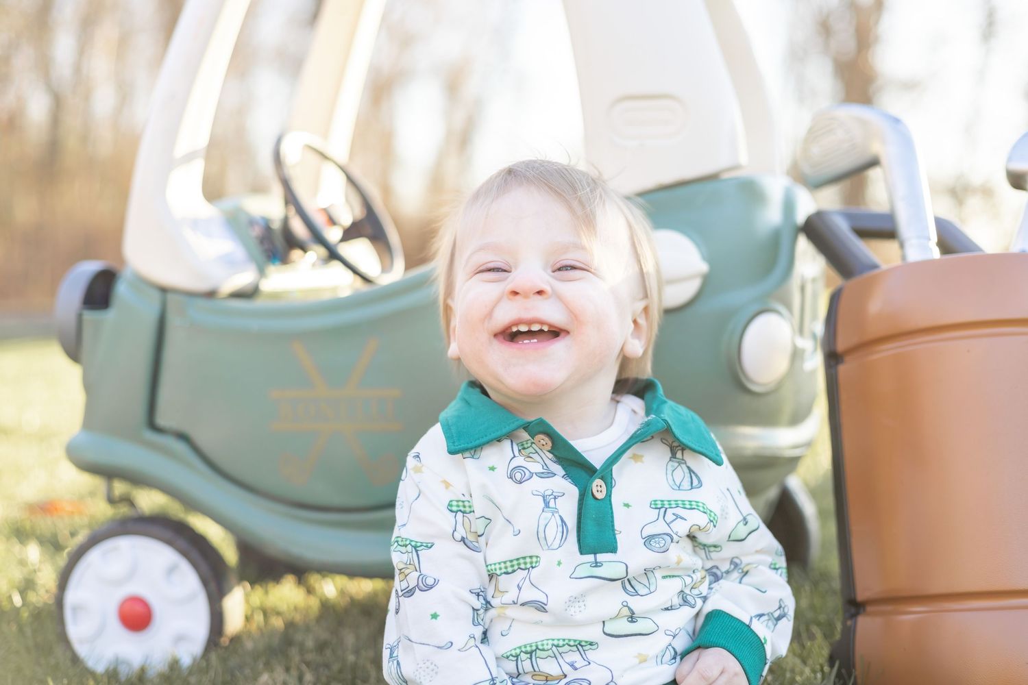 A child in a patterned shirt sits outdoors with a big joyful smile, with a vintage wagon wagon in the background.