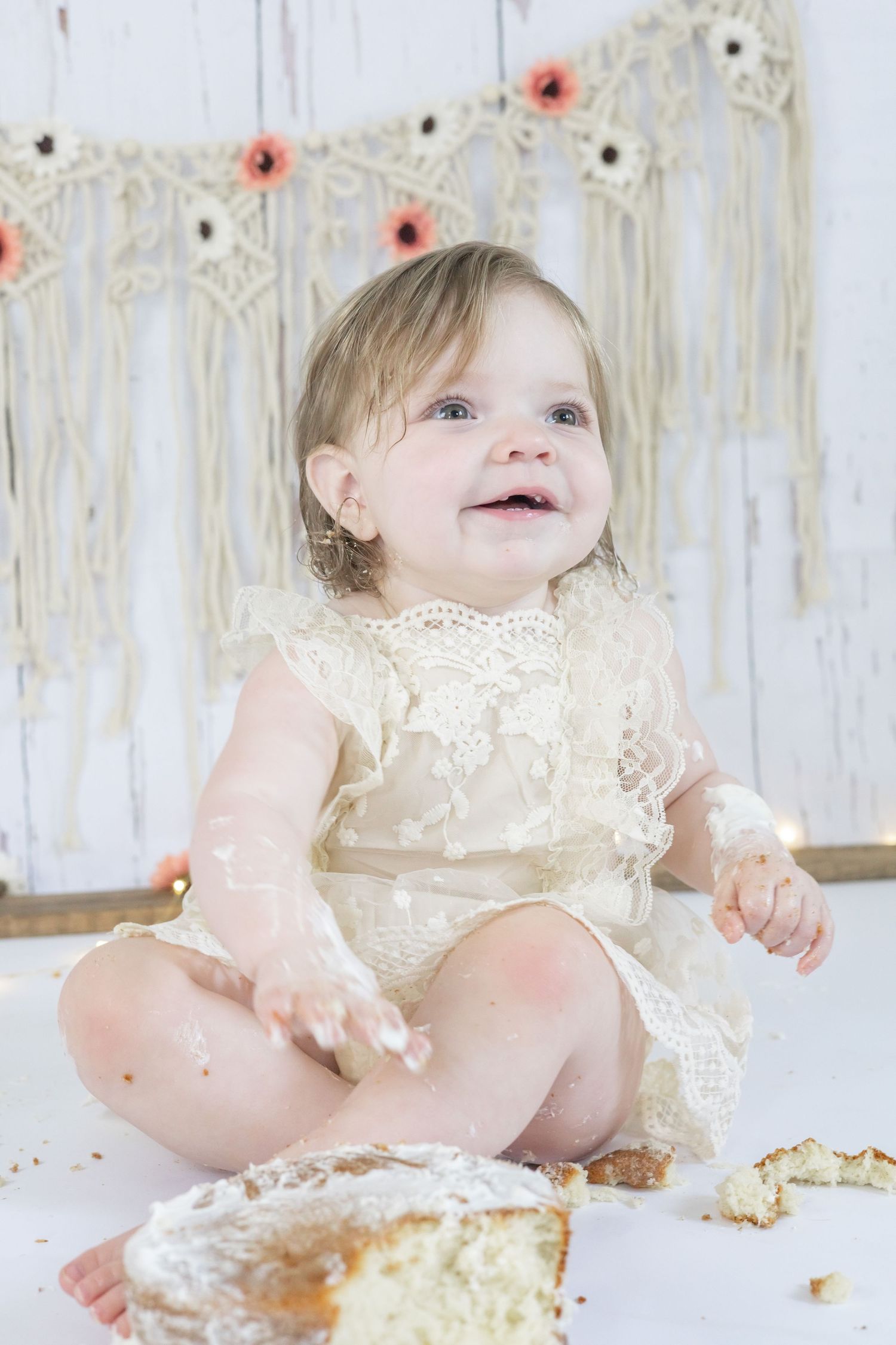 A small child in a cream lace dress sits on a white blanket with cake smashed around them during a cake smash photo session.