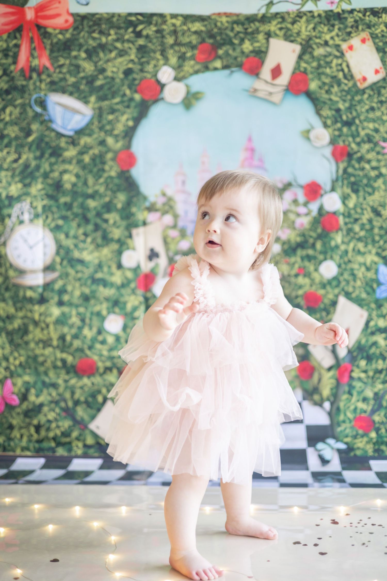 Baby in pink tulle dress plays on checkered floor with fairy lights and tea party decorations in background.