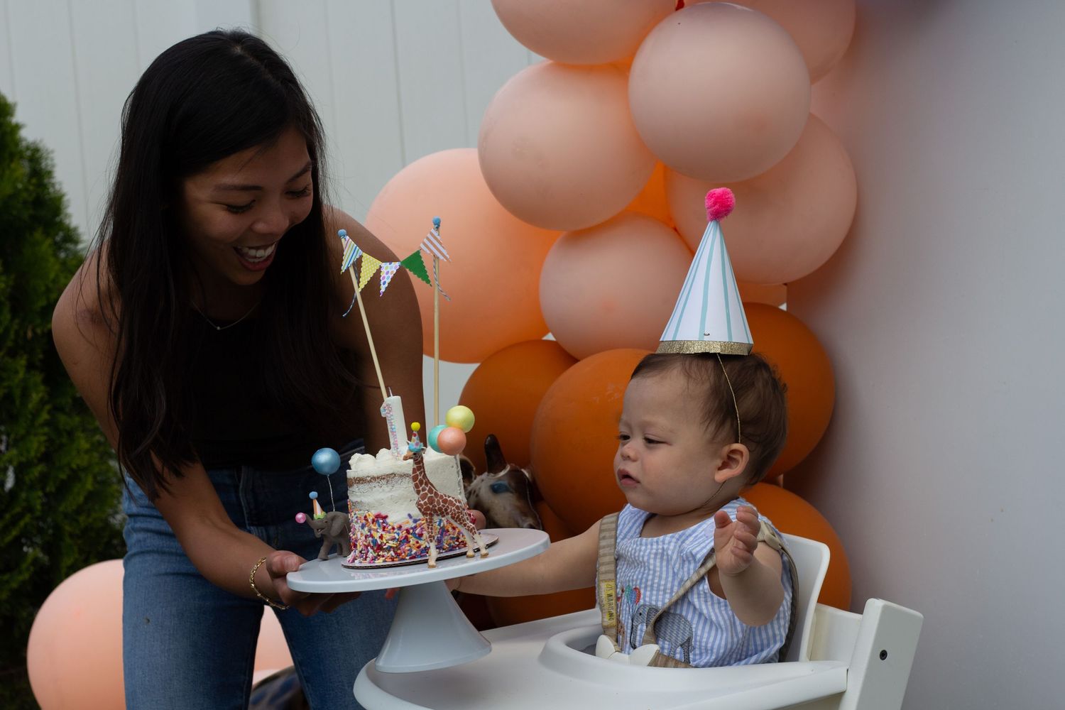 A baby in a high chair celebrates their first birthday with a cake and peach-colored balloon decorations.