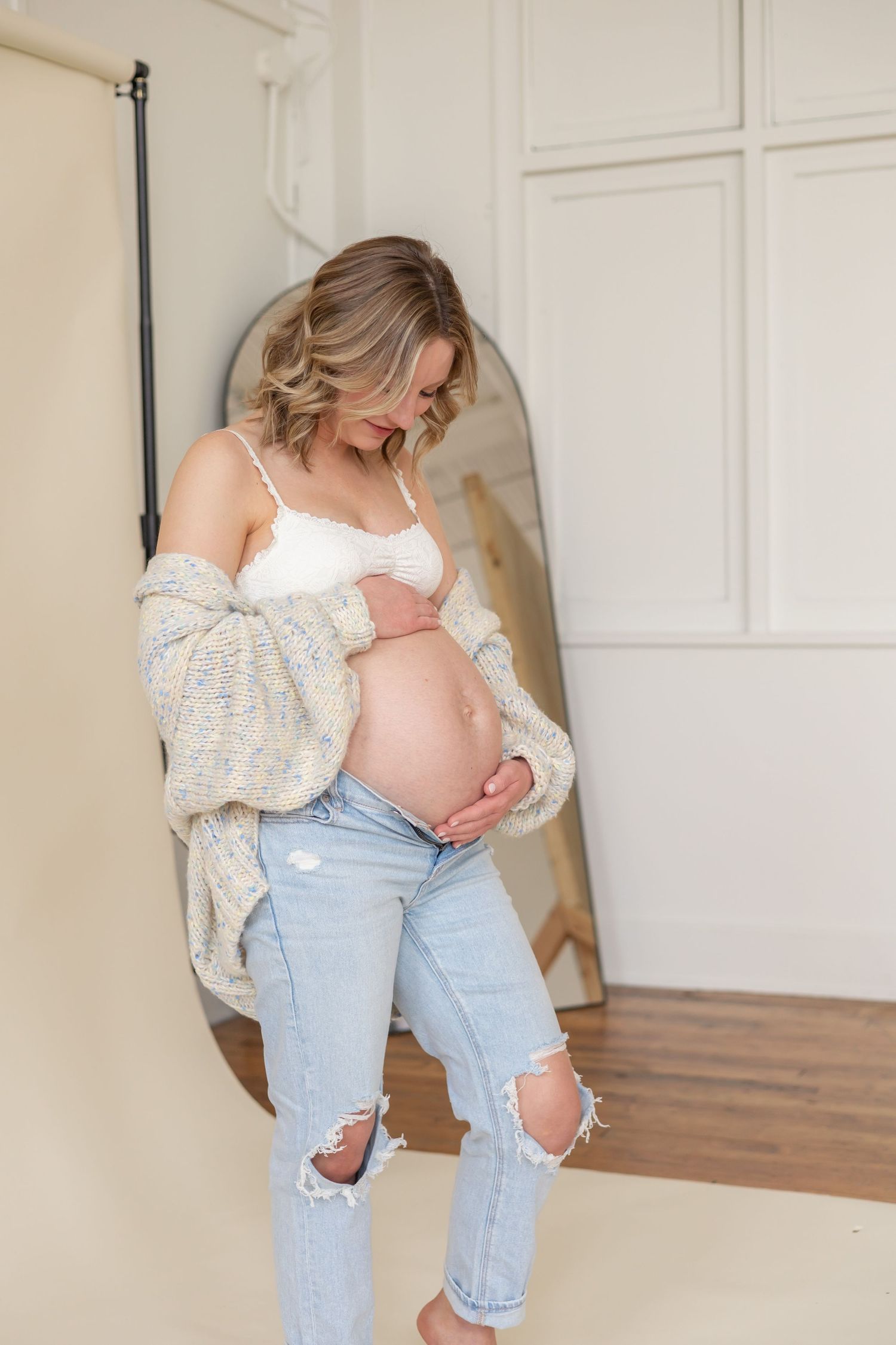Expectant mother in ripped jeans and cozy sweater poses against white wall during indoor maternity photoshoot.