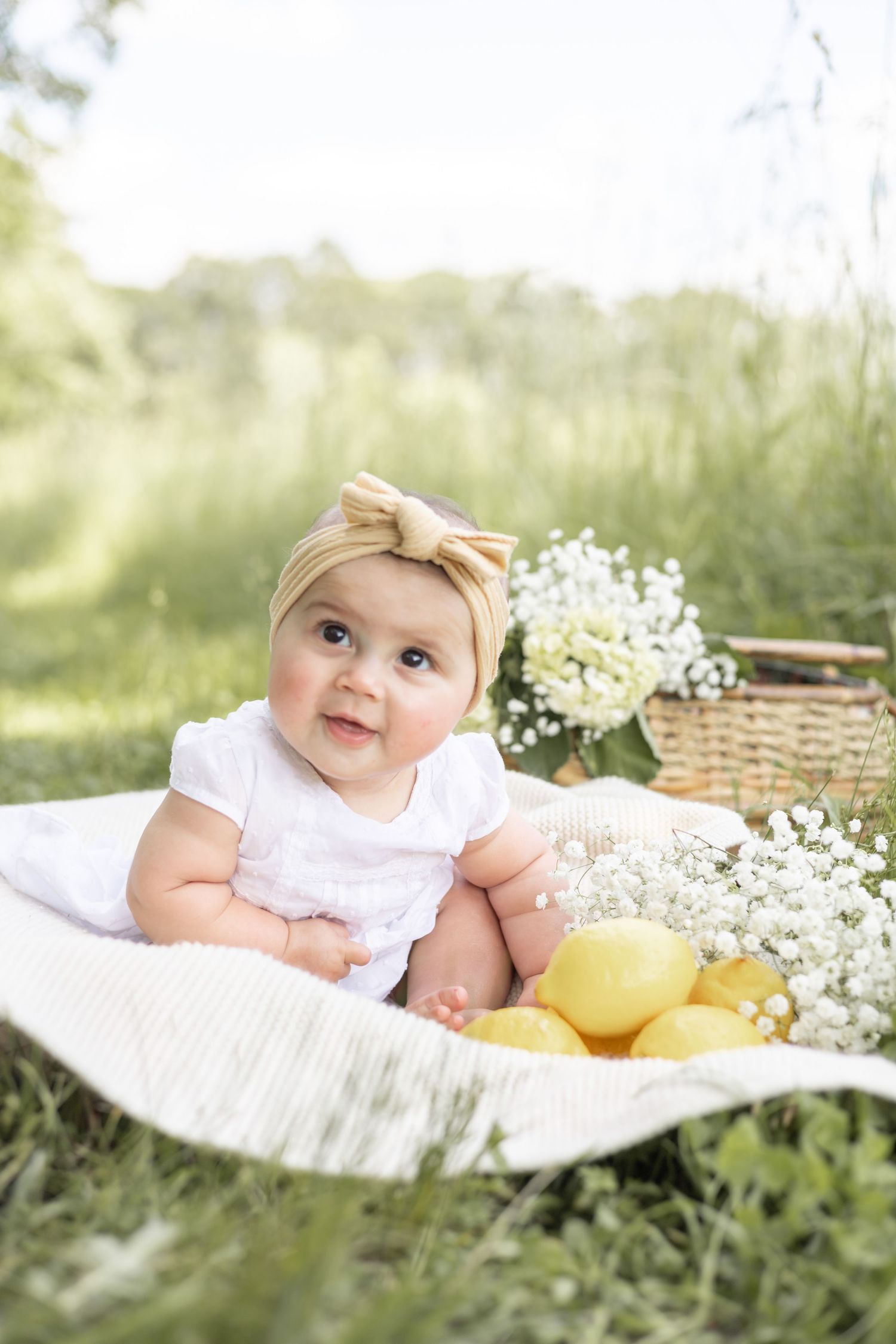 Baby in white dress sits on blanket outdoors with lemons and baby's breath flowers in natural summer setting.