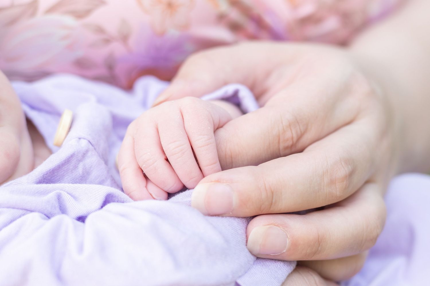A tiny baby's hand gently holds onto an adult's finger while resting on soft purple fabric.