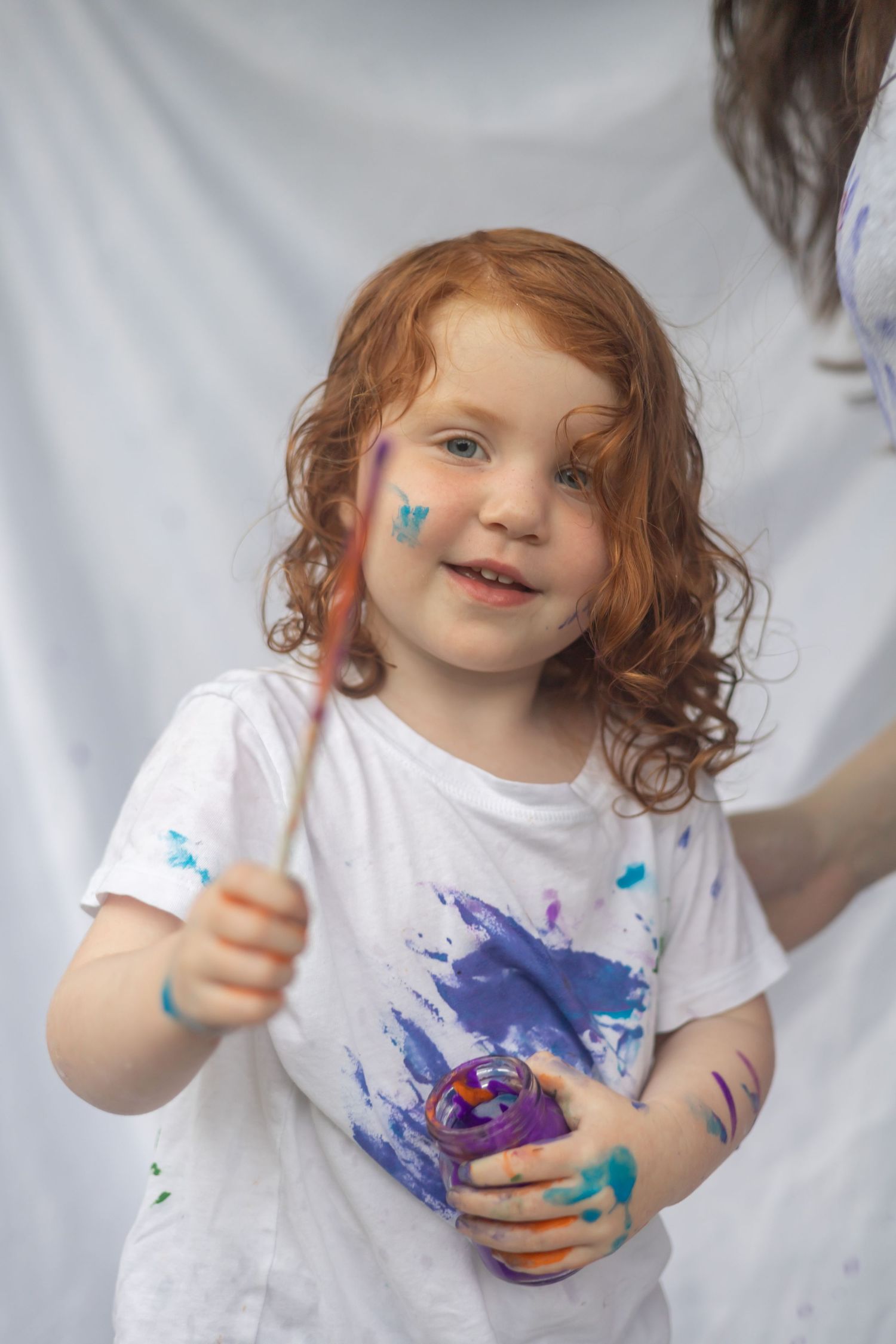Child wearing white t-shirt covered in blue paint splatters holds art supplies with a happy expression.