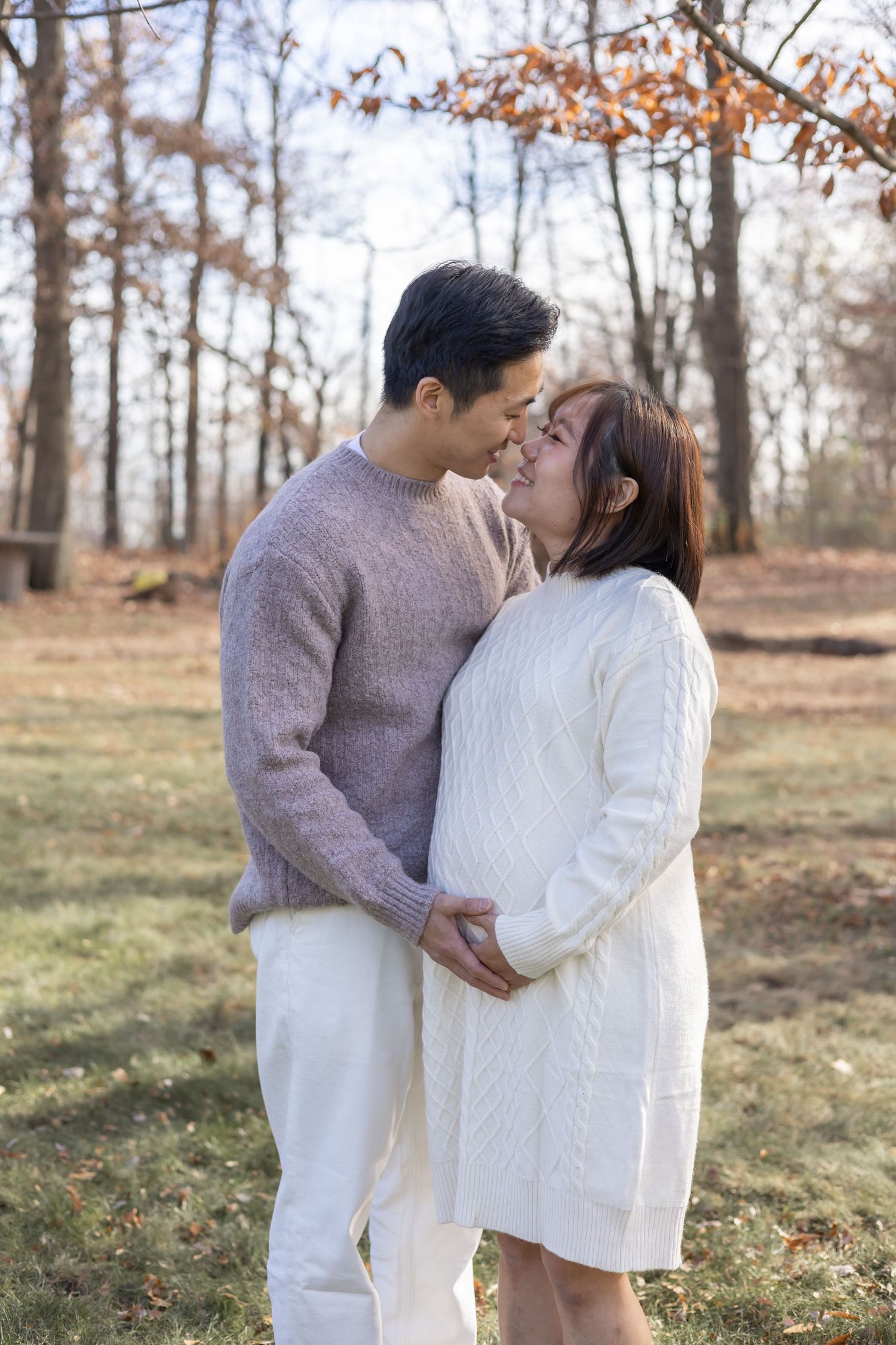 A romantic autumn scene showing a couple embracing in a wooded area wearing matching cream and gray sweater outfits.