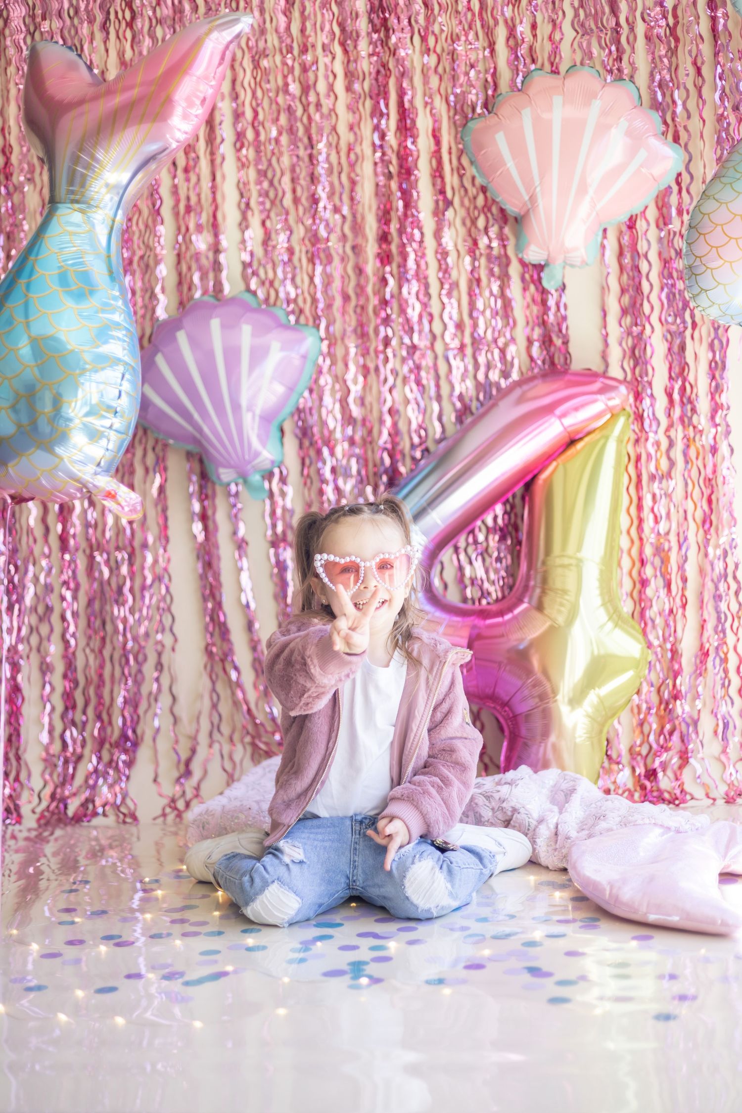 A dreamy pastel party setup with seashell balloons, pink tinsel backdrop, and iridescent confetti on the floor.