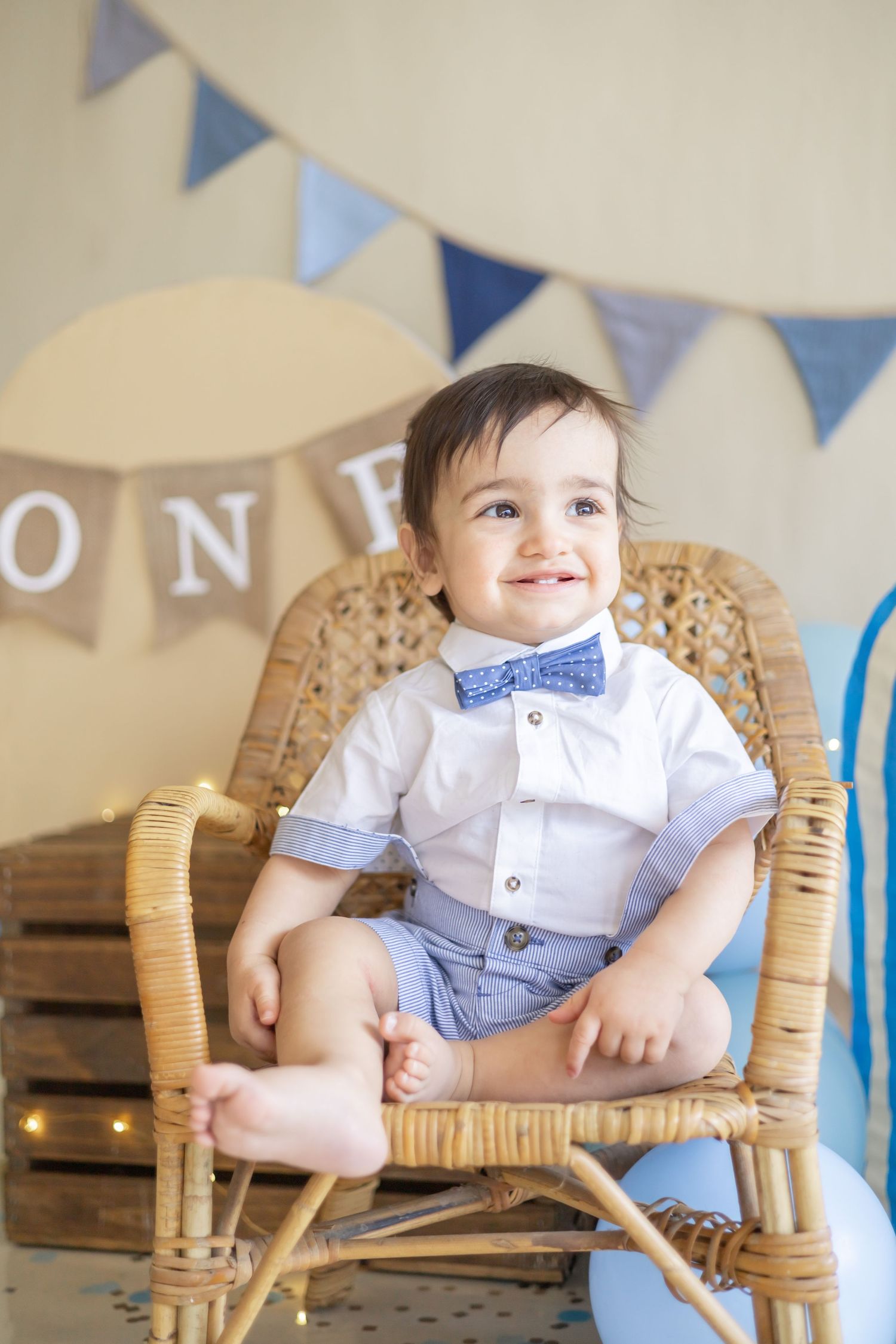 Baby wearing white shirt and blue bow tie sits in wicker chair with blue banner decorations in background.