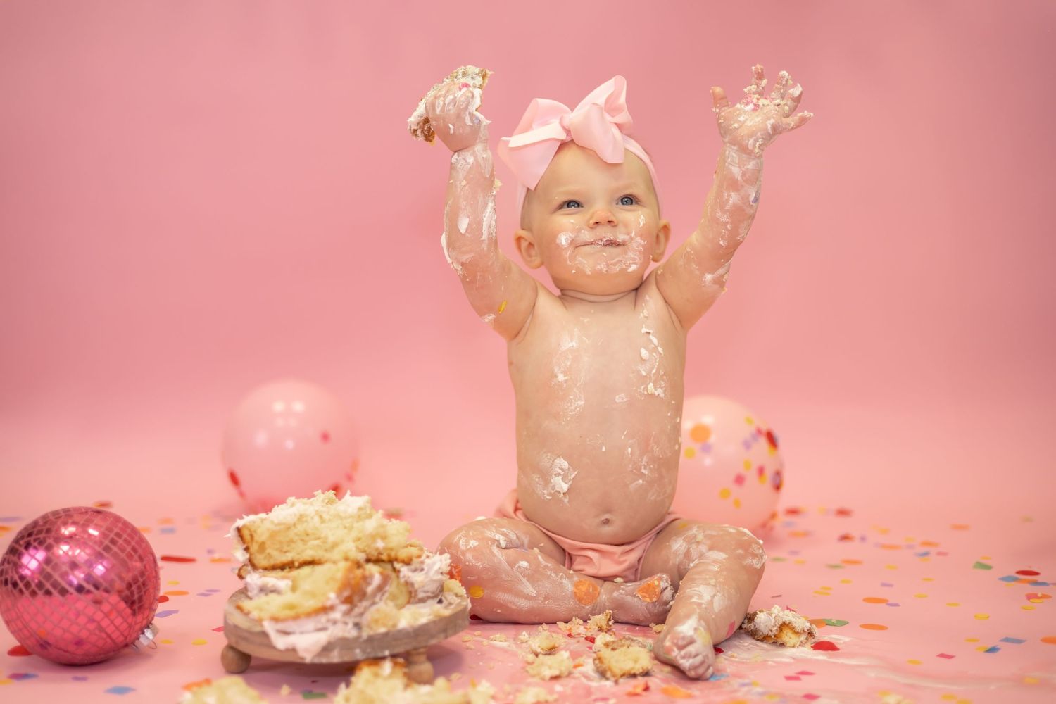 Baby covered in cake and confetti during pink-themed cake smash photo session with balloons and decorations.