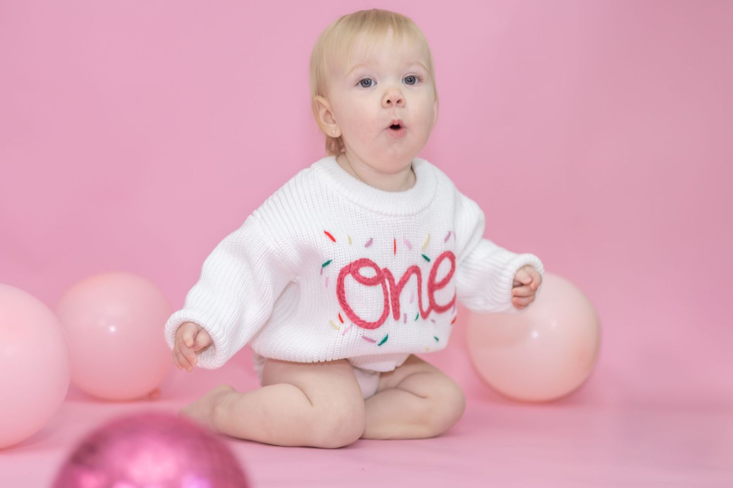 Baby in white birthday outfit sitting among pink balloons during first birthday photoshoot.