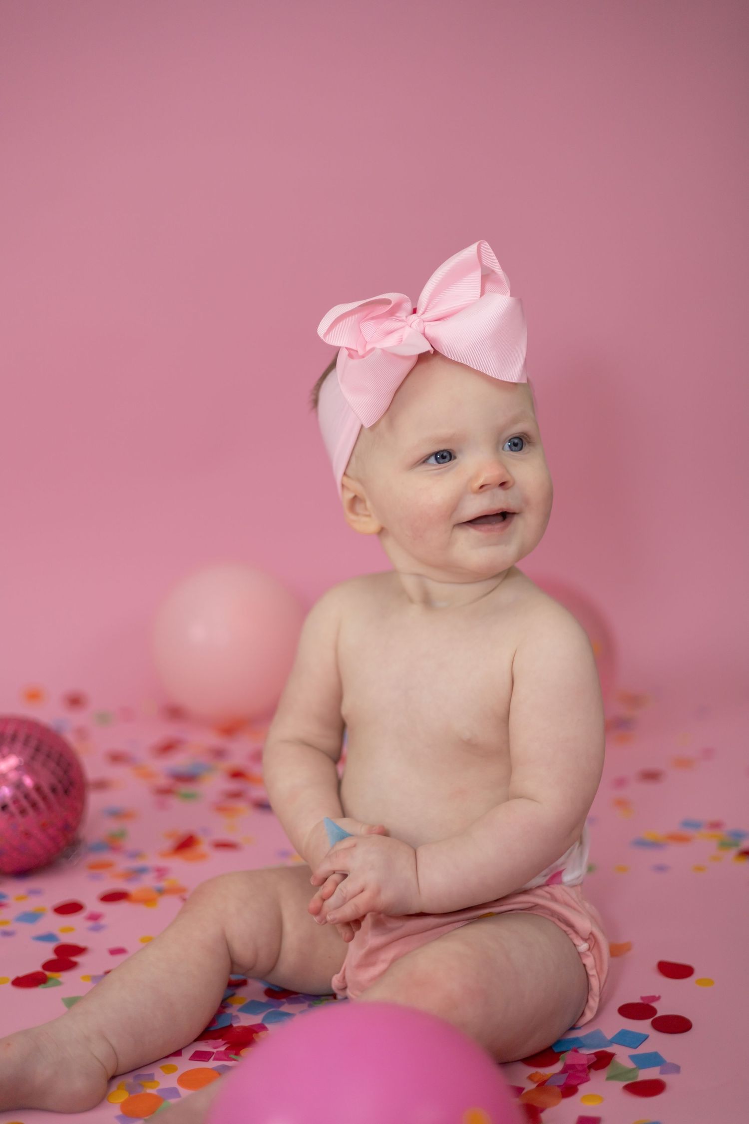 Baby in pink bow headband sitting on floor with pink balloons and confetti for birthday celebration.