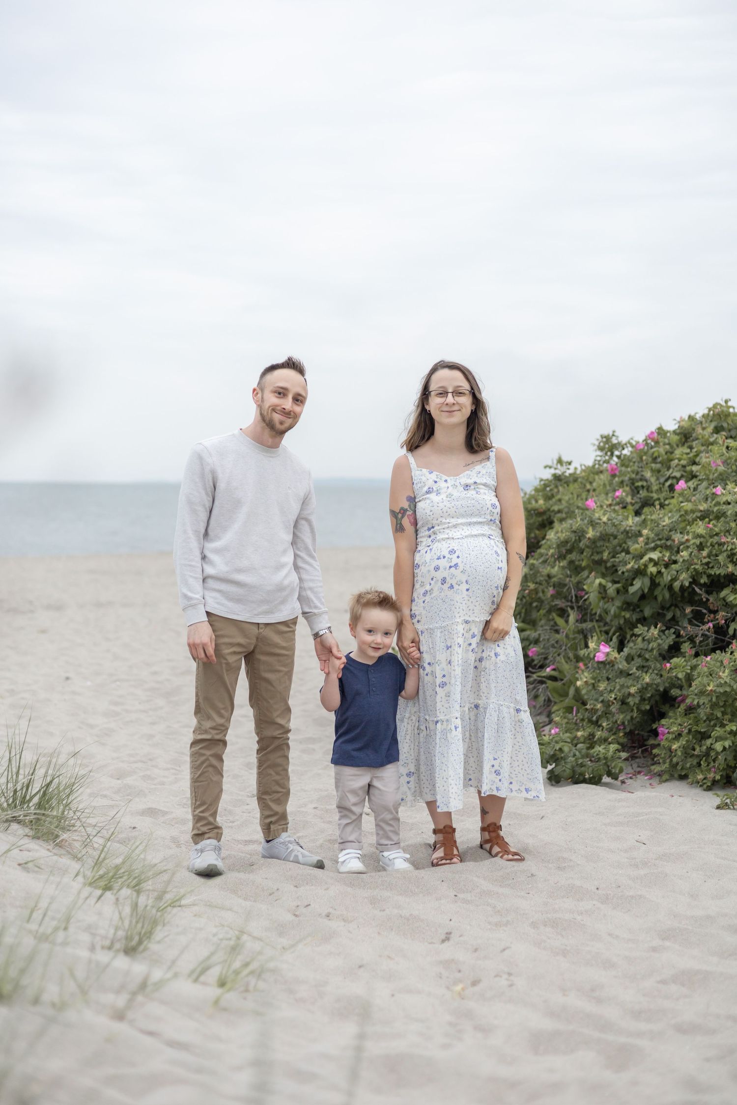 A family walks together on a sandy beach near green coastal vegetation on a cloudy day.