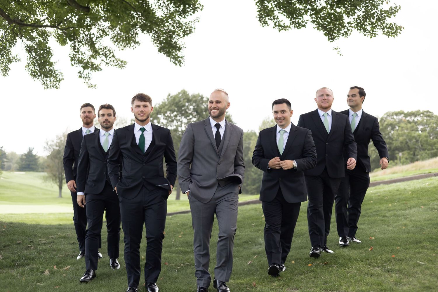 Groomsmen in dark suits walking together across a grassy hillside at an outdoor wedding venue.