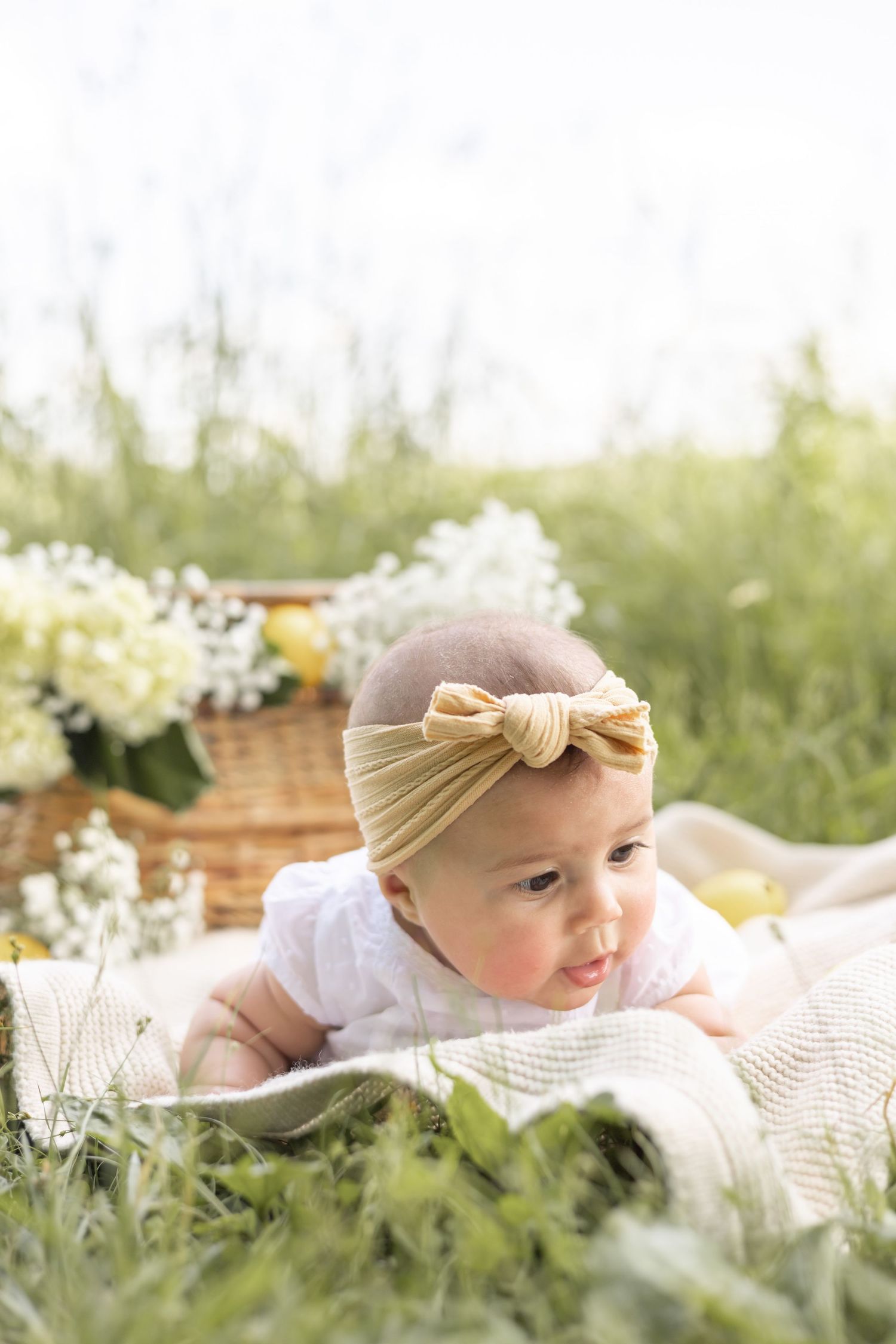 Baby in cream headband lying on blanket outdoors surrounded by white flowers and greenery in sunlit garden setting.