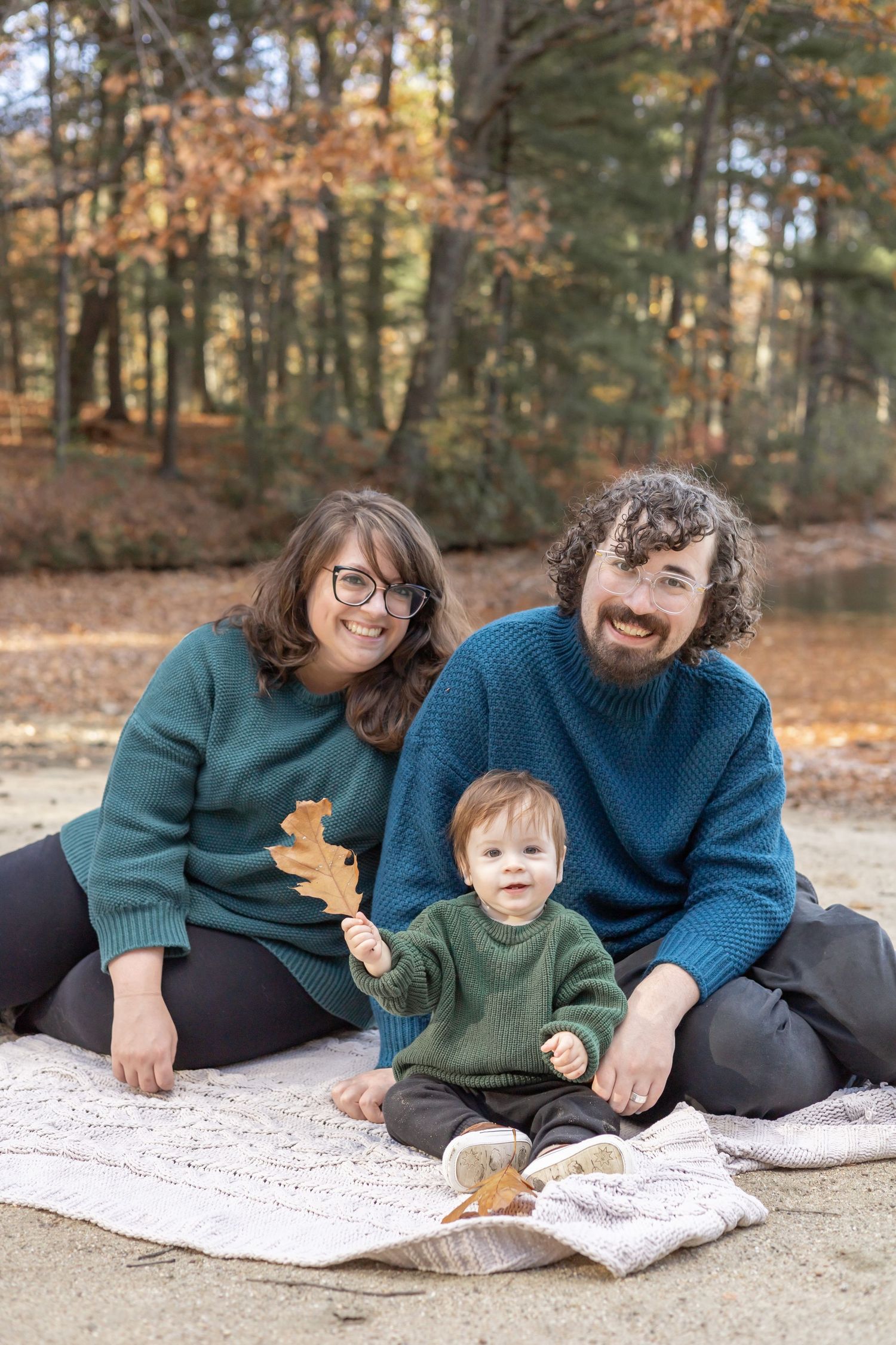 A family in matching blue and green sweaters sits on a blanket surrounded by autumn leaves in a forest setting.