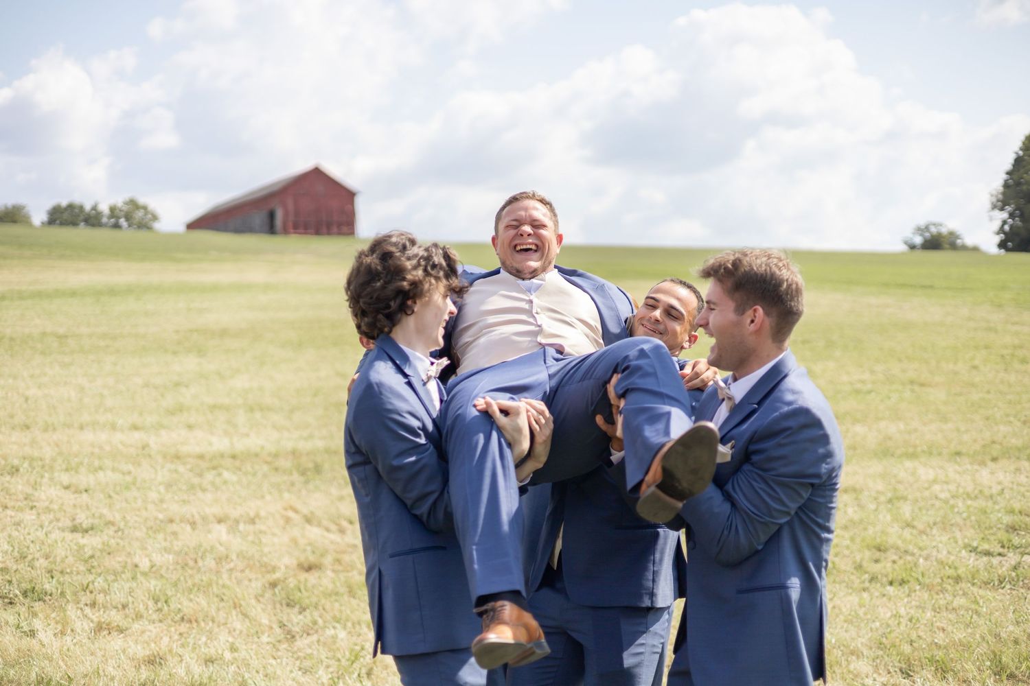 Celebratory moment on grassy field with three people in blue suits lifting someone in front of a red barn.