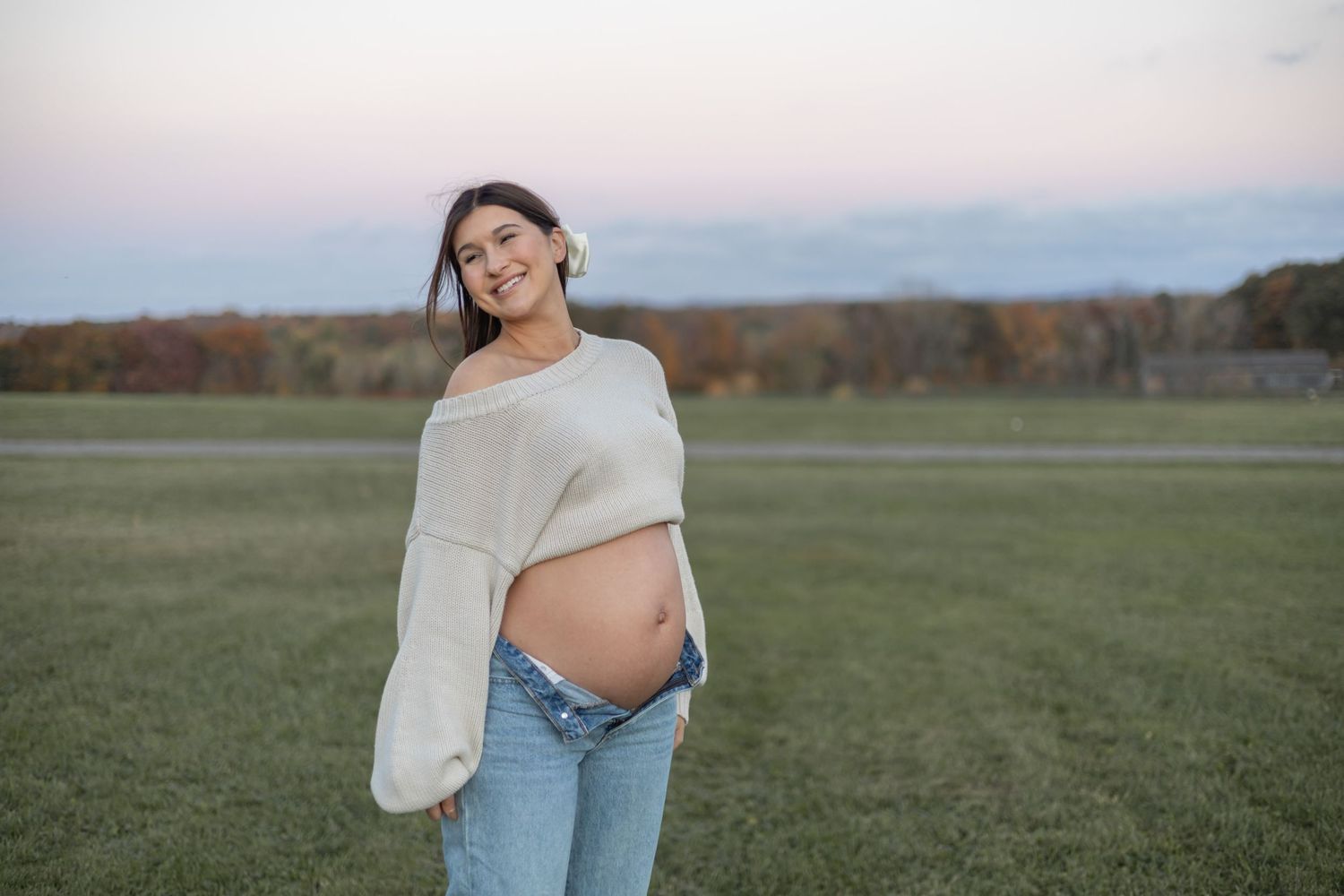 Maternity photo of pregnant figure wearing white sweater and jeans while standing in open field at dusk.