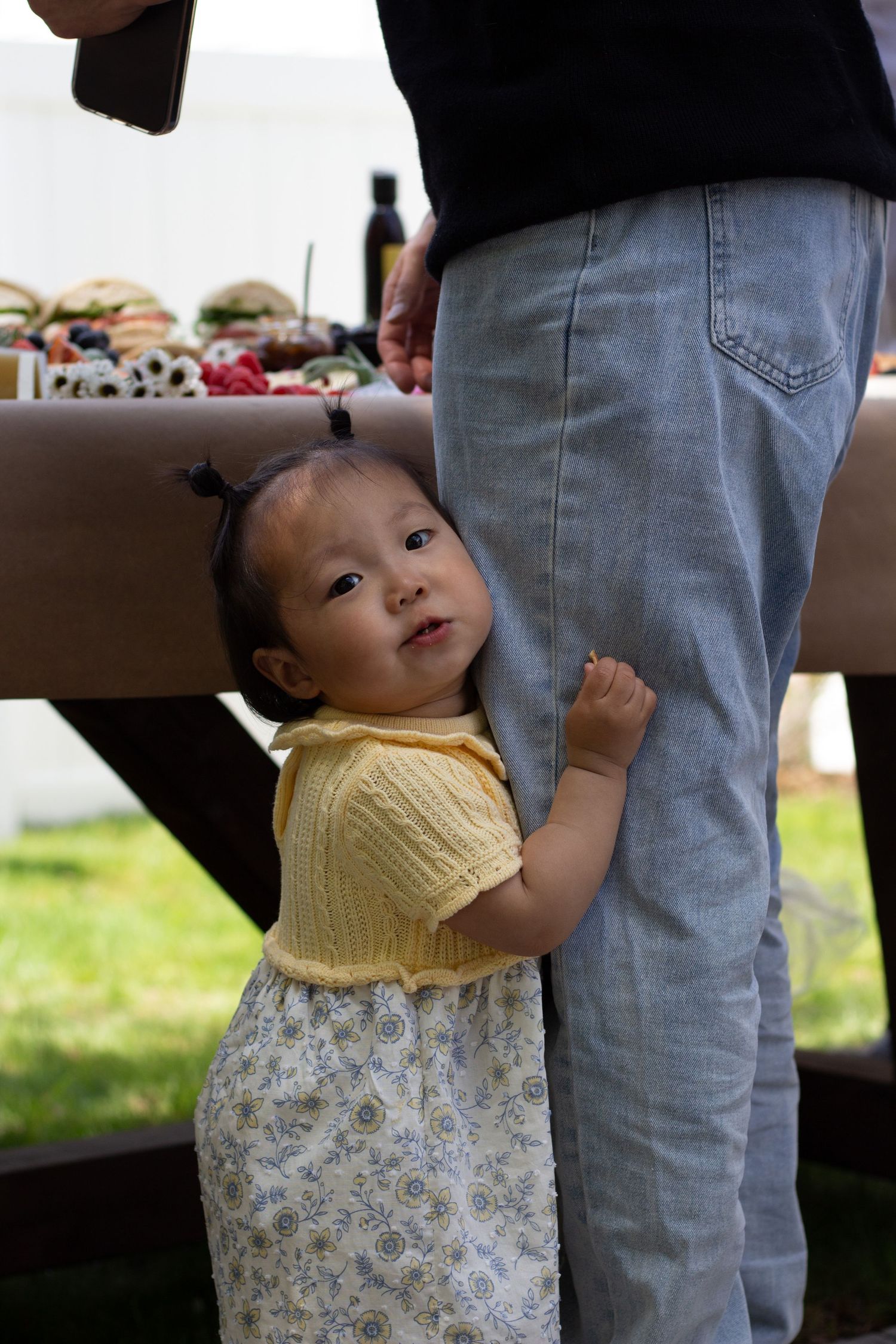 A young child in a yellow top and floral dress holds onto someone's leg while standing on grass outdoors.