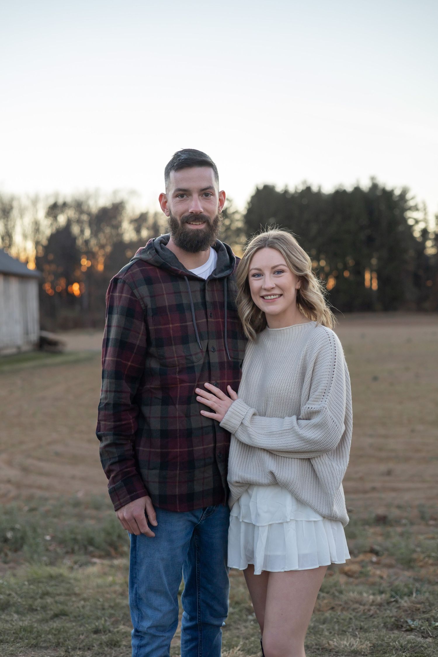 Couple in casual clothing posing together at sunset in a rustic outdoor setting.