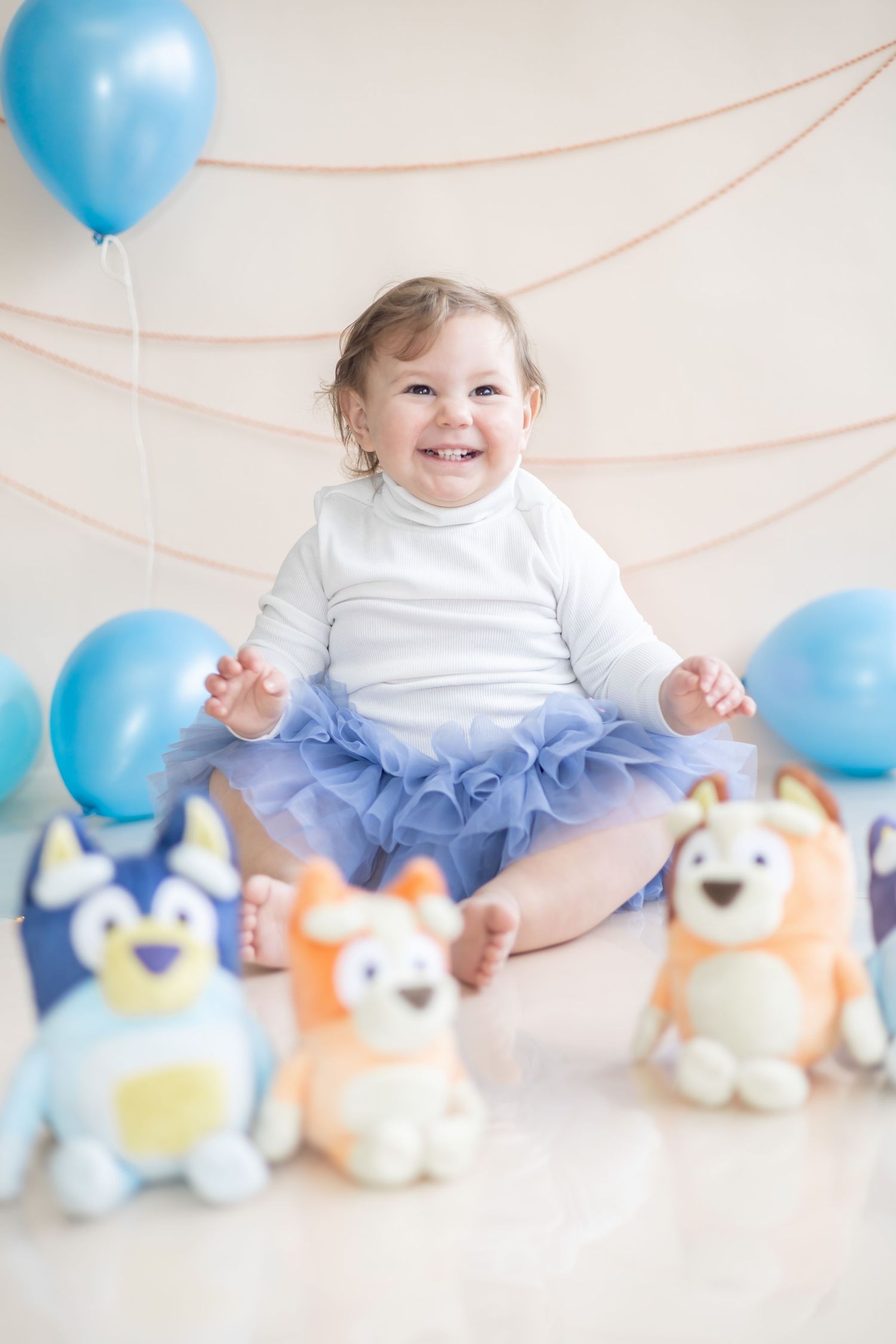 Baby in white top and tulle skirt sits on floor with blue balloons and plush animal toys in studio setting.