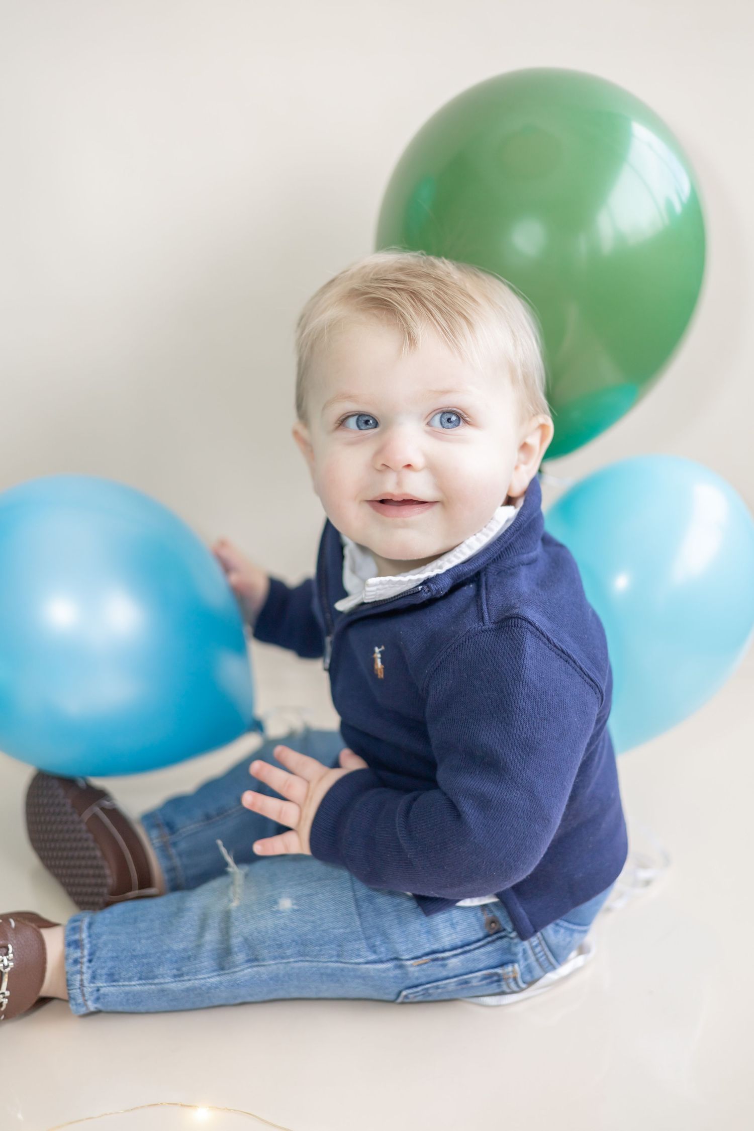 Small child in navy sweater playing with blue and green balloons on light background while sitting on floor.