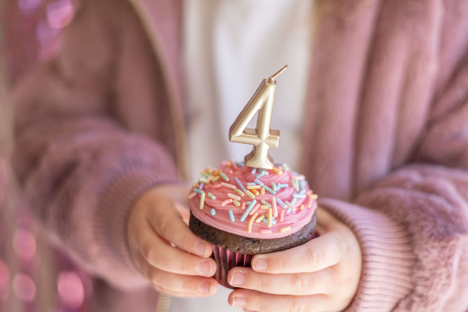 Hands hold a chocolate cupcake with pink frosting and rainbow sprinkles topped with a gold number 4 candle.