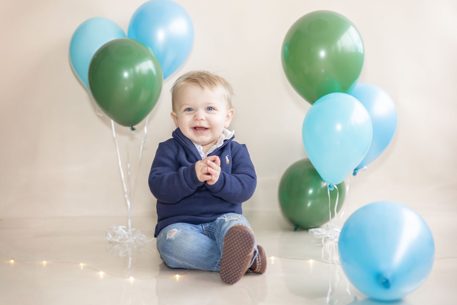 A baby sits on a white floor surrounded by blue and green balloons during a photo session.