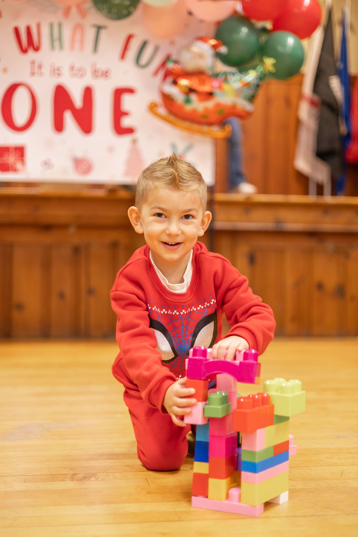 A young child in a red sweater playing with colorful building blocks on a wooden floor during a celebration.