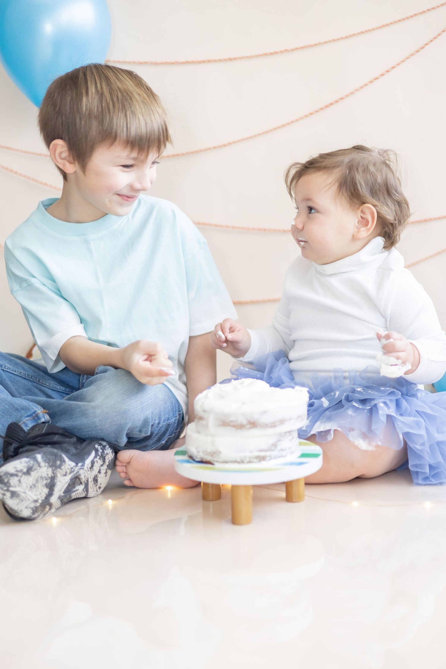 Two children in blue and white clothes share dessert and interact playfully against a soft-colored background.